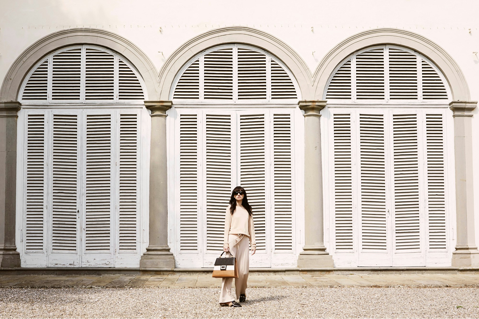 Image shows a person wearing an off-white top and pants, holding a striped leather bag. They are standing in front of 3 arches filled with slatted shutters.