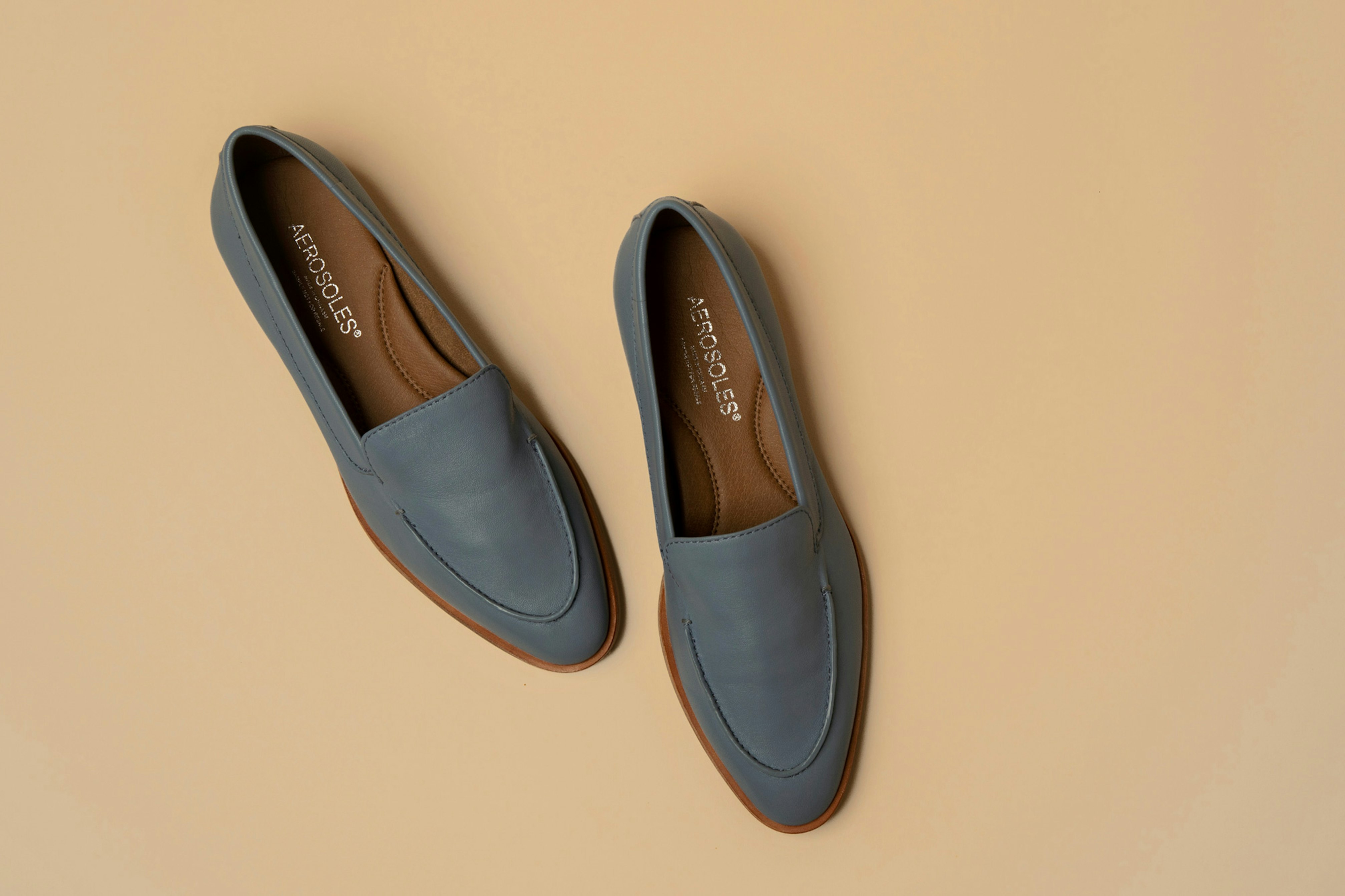 Image showing overhead shot of a pair of loafers in a teal color placed on a beige seamless. The logo is sowing in gold inside the shoes.