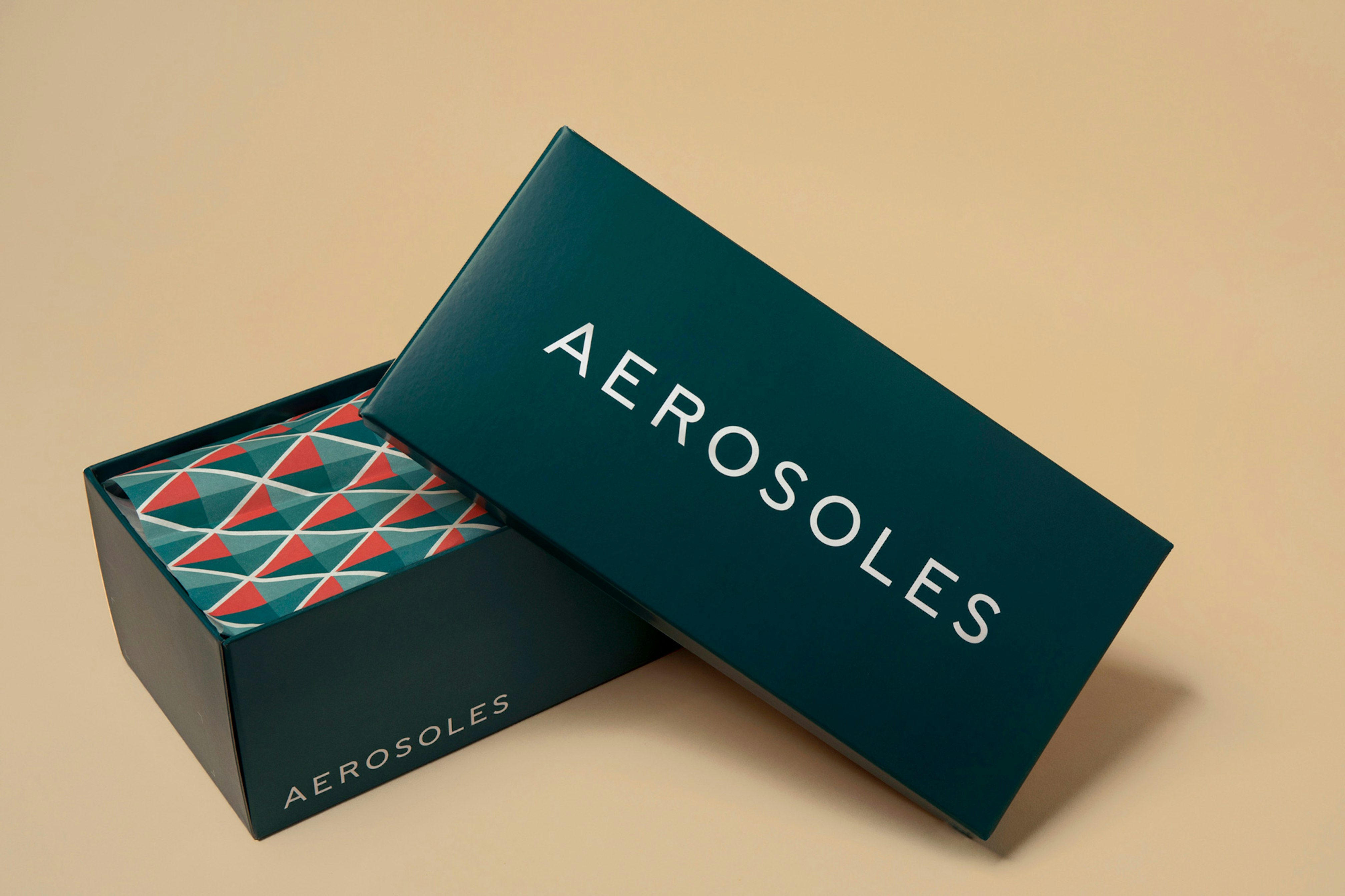 Image showing the teal colored shoe box half open revealing a colorful diamond shaped tissue paper inside. The logo is written across the lid in white.