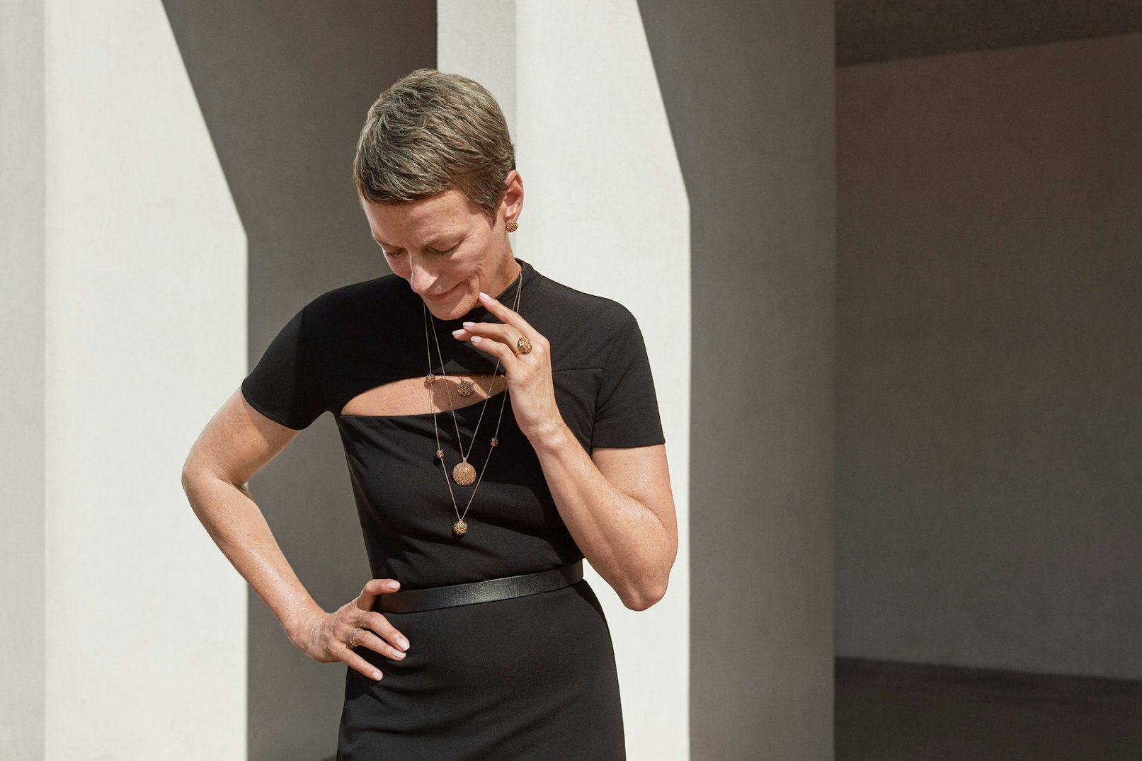 Person standing outside against architectural details. Person is wearing a black dress and necklaces.