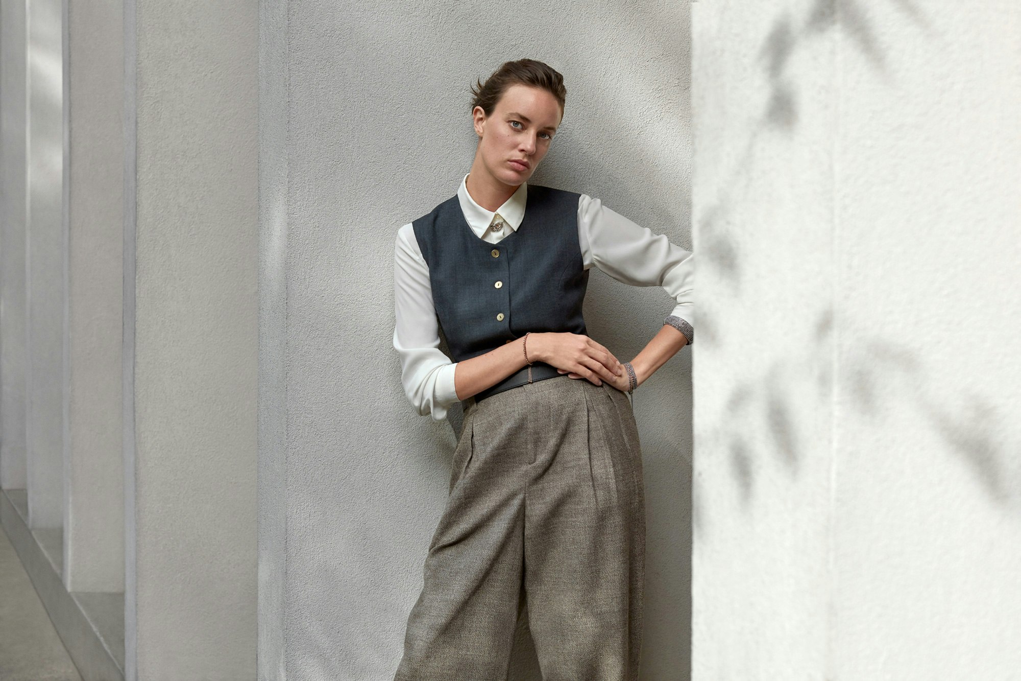 Photo showing person leaning on a white outdoors wall. There are plant shadows on the wall and the person is wearing oversized tweed pants, a light gray blouse, and a dark teal vest on top.