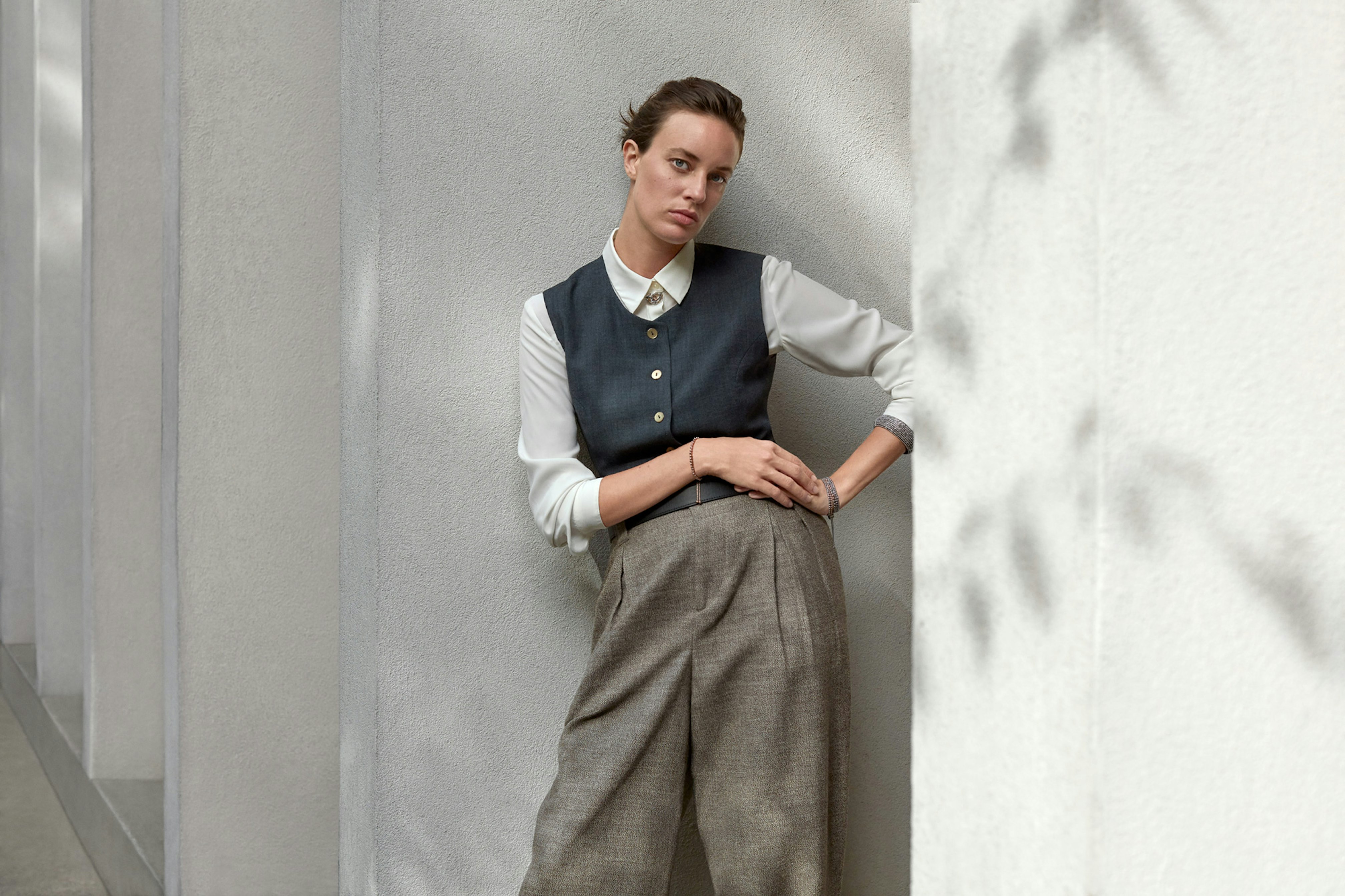 Photo showing person leaning on a white outdoors wall. There are plant shadows on the wall and the person is wearing oversized tweed pants, a light gray blouse, and a dark teal vest on top.