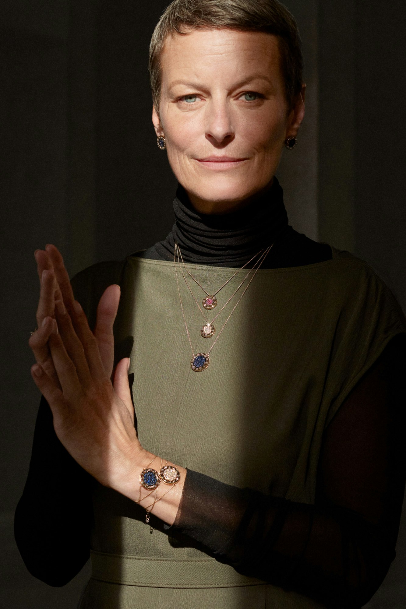 Photo showing person looking in camera. They are wearing an olive green dress on top of a black turtleneck. Three necklaces are layered on top of the dress.