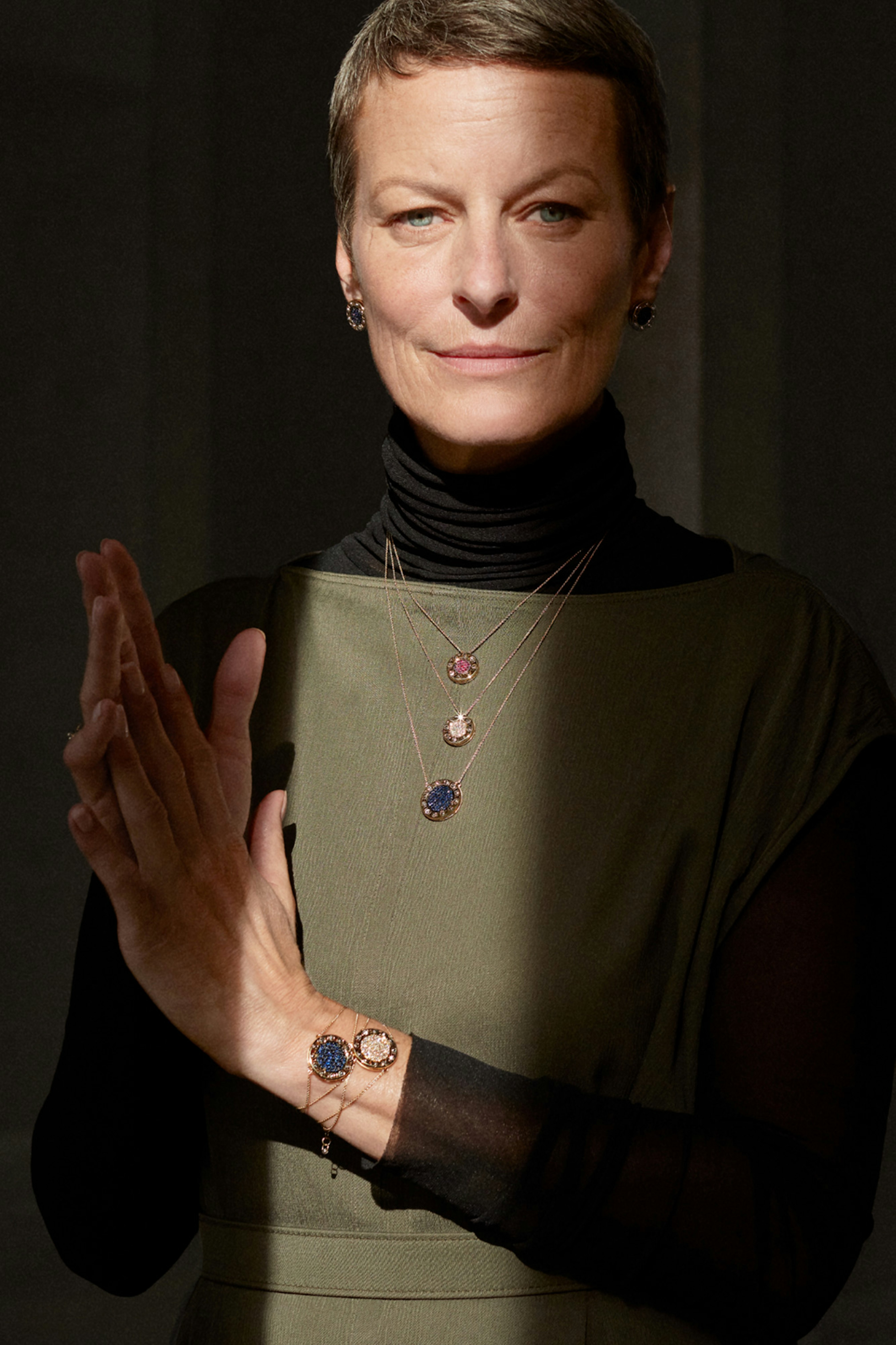 Photo showing person looking in camera. They are wearing an olive green dress on top of a black turtleneck. Three necklaces are layered on top of the dress.