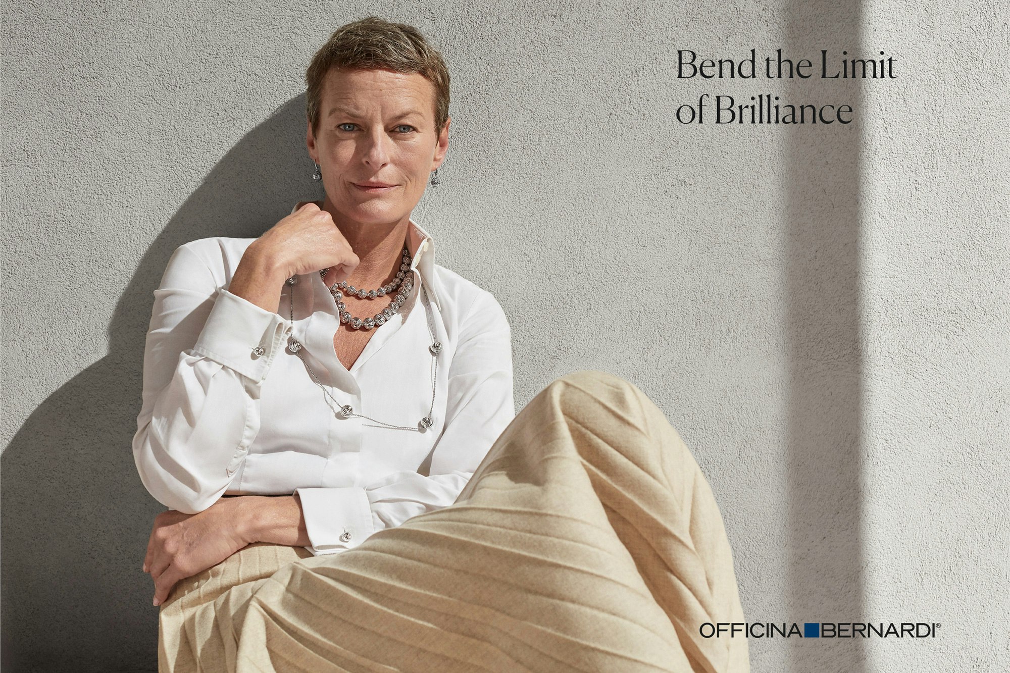 Image showing person seated outside against a concrete wall. They are looking in camera, have a pixie haircut and are wearing a white crisp shirt and a cream colored pleated long skirt.