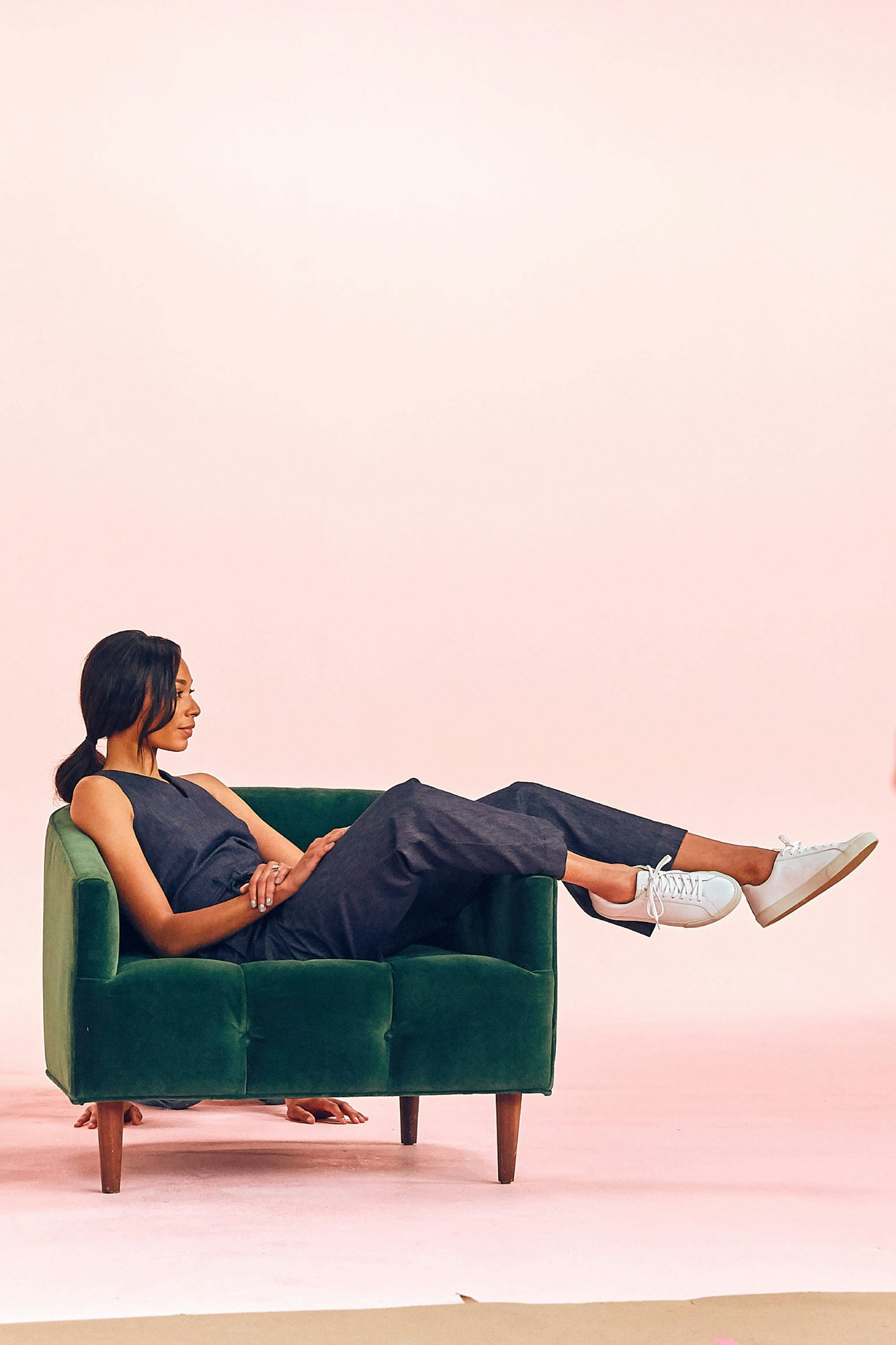 Image showing person sitting in a green chair. They are wearing a denim jumpsuit and white sneakers and the seamless is petal pink.