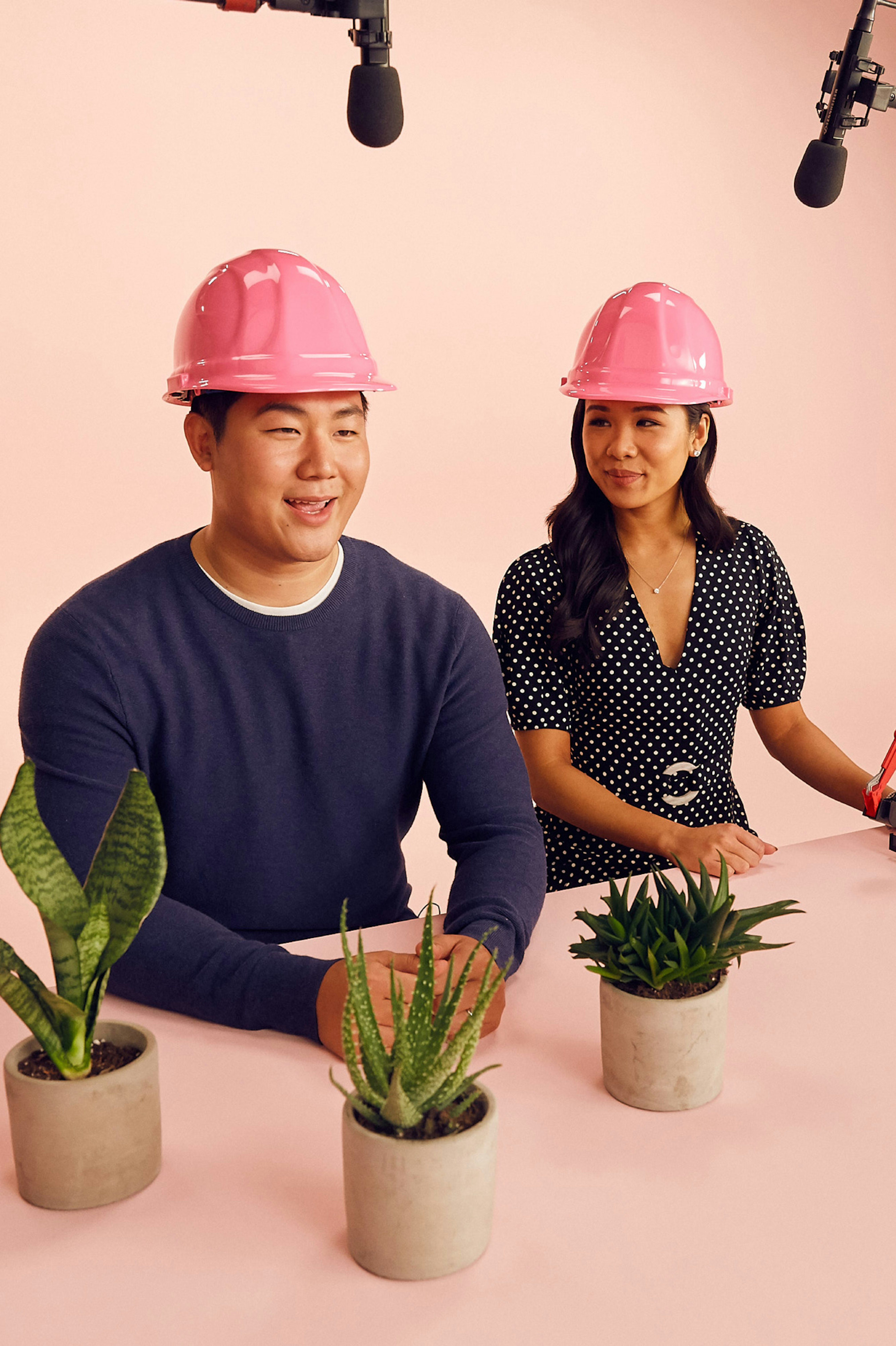 Image showing two people at a table wearing pink hard hats. The table and the seamless are soft pink and microphones are showing. On the table are three green small plants in gray vases.
