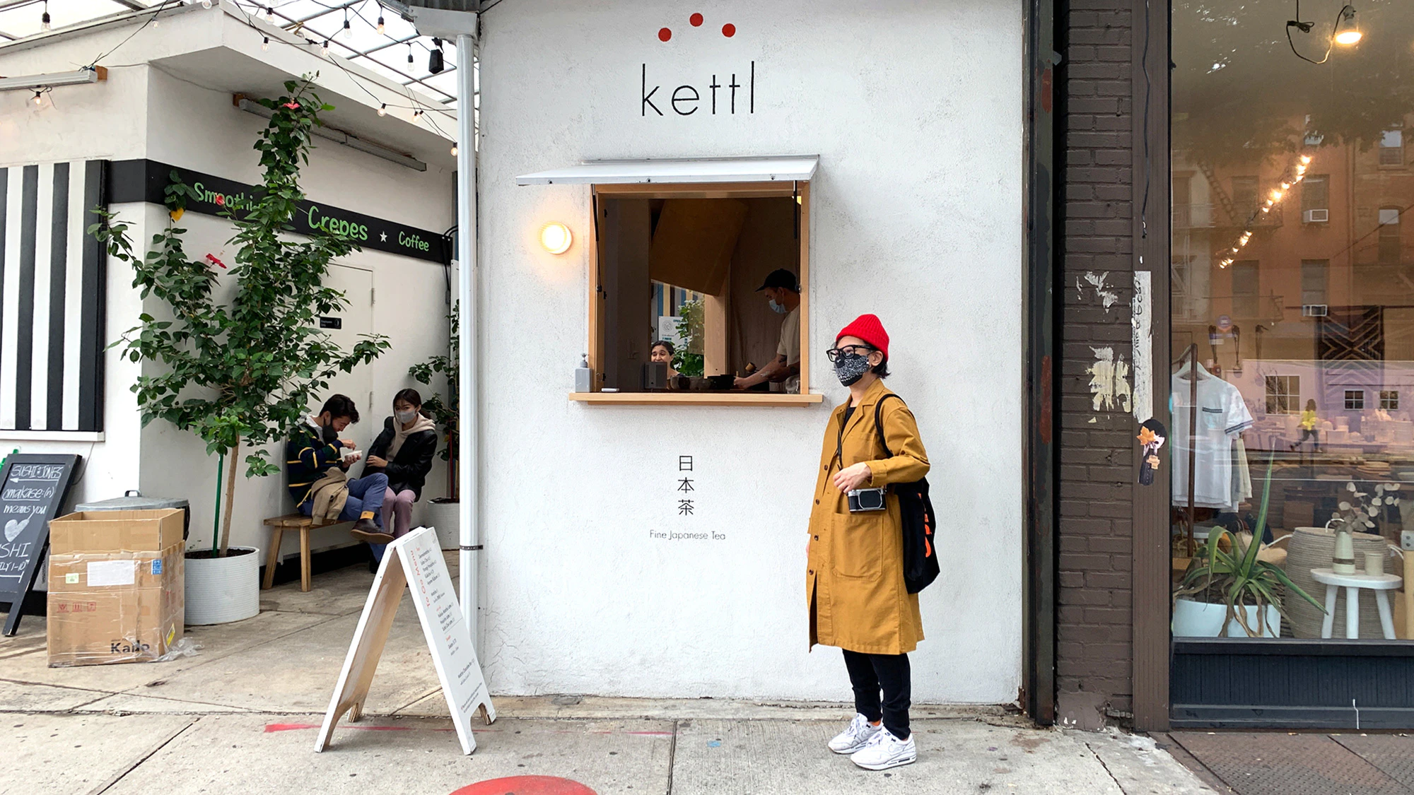 Image showing person standing outside the Kettl cafe. They are wearing a red hat and a medium brown long jacket. The cafe wall is white with the logo on and Japanese writing.