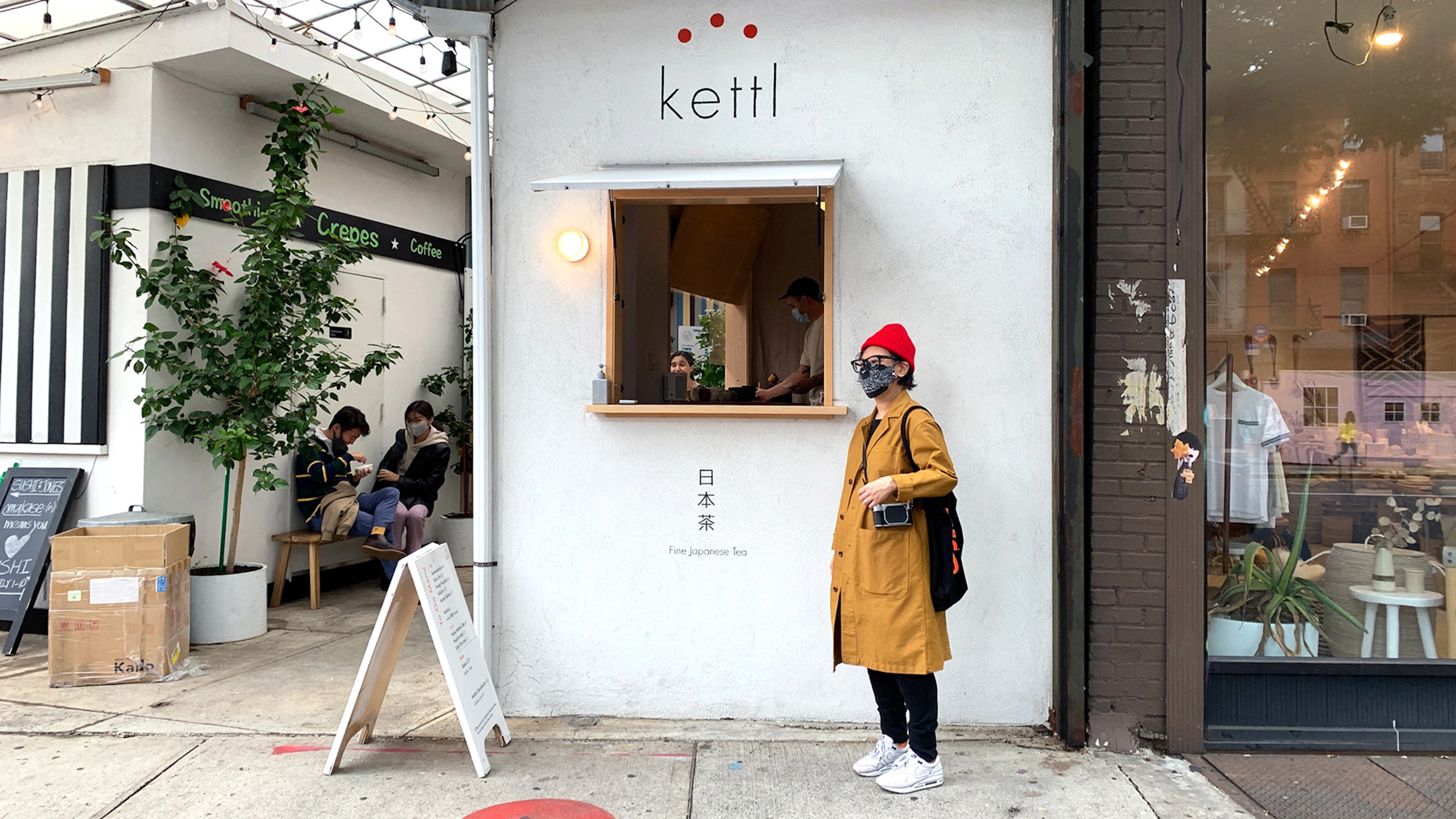 Image showing person standing outside the Kettl cafe. They are wearing a red hat and a medium brown long jacket. The cafe wall is white with the logo on and Japanese writing.