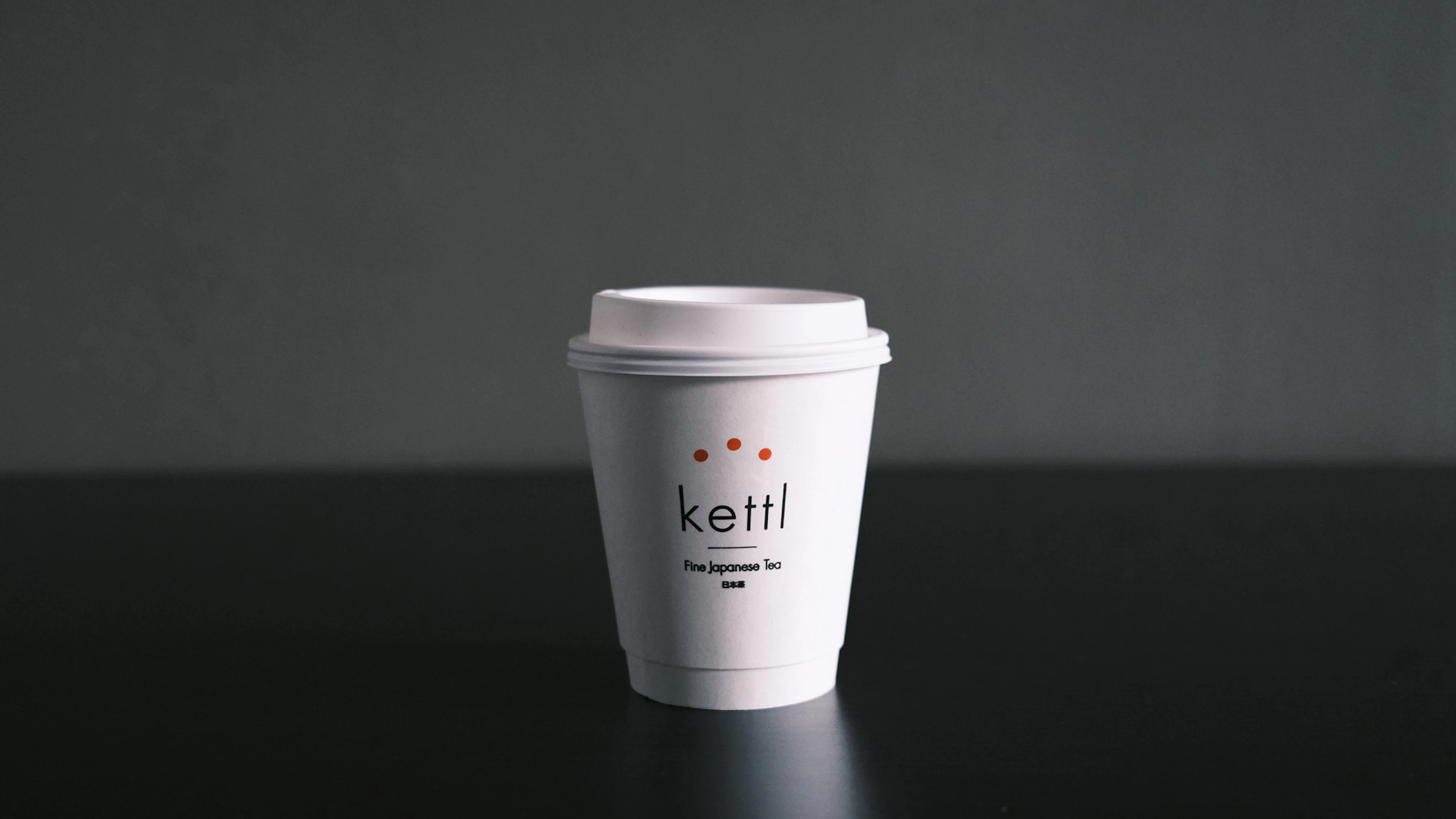 Image showing a white take-out cup with Kettl logo on the front and a white plastic lid. The table is black and the wall dark gray.