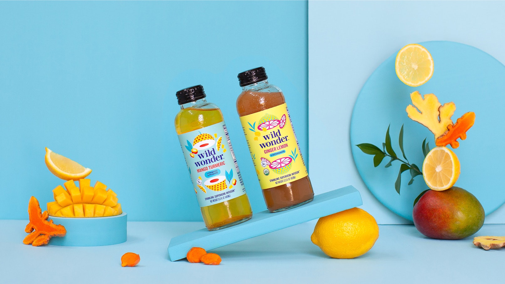 Two glass bottles filled with juice are sitting at an angle on a blue board. The seamless is blue as well and around the bottles are mangoes, lemons, ginger, and turmeric root.