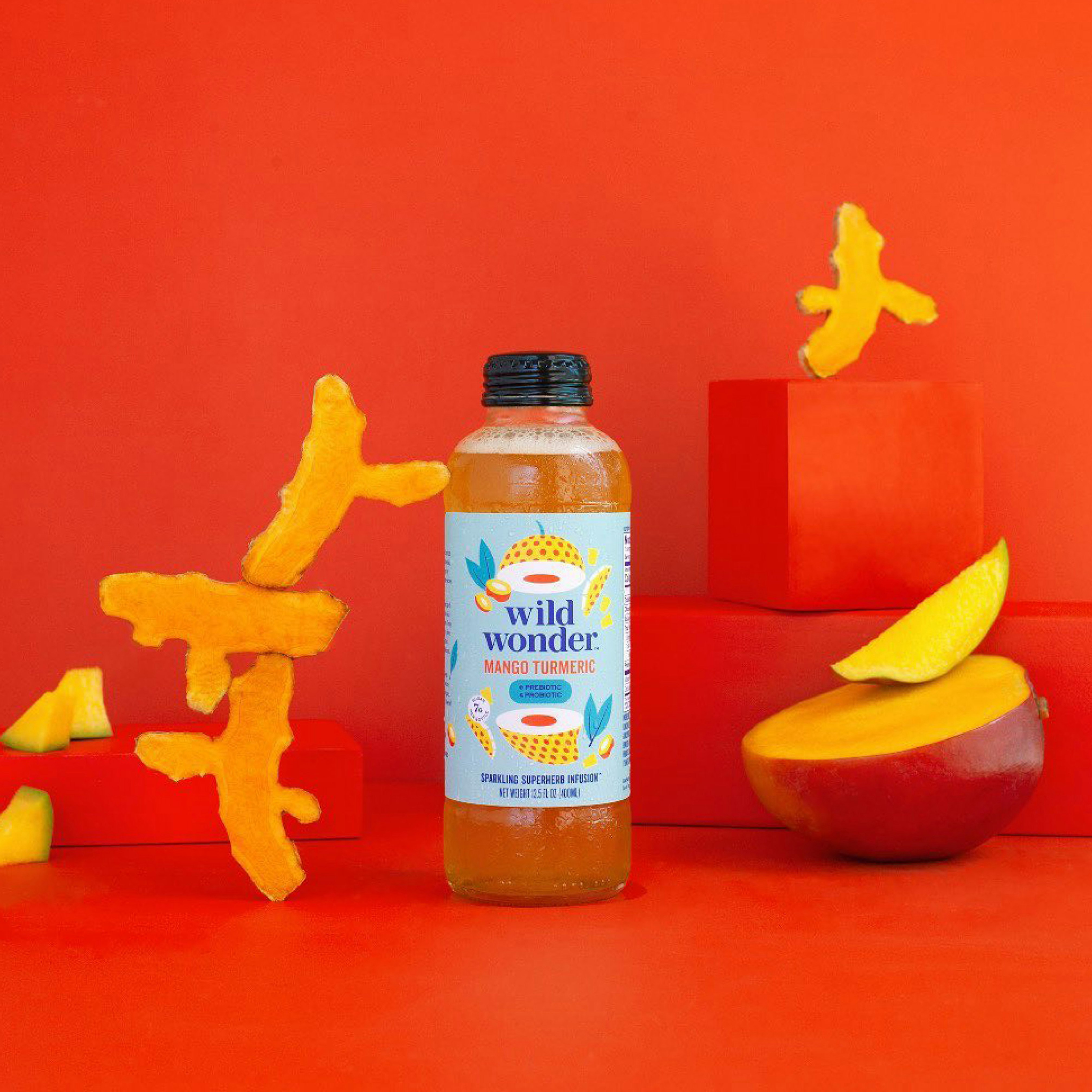 Image of bottle standing on a red seamless with turmeric root and mango slices next to it.