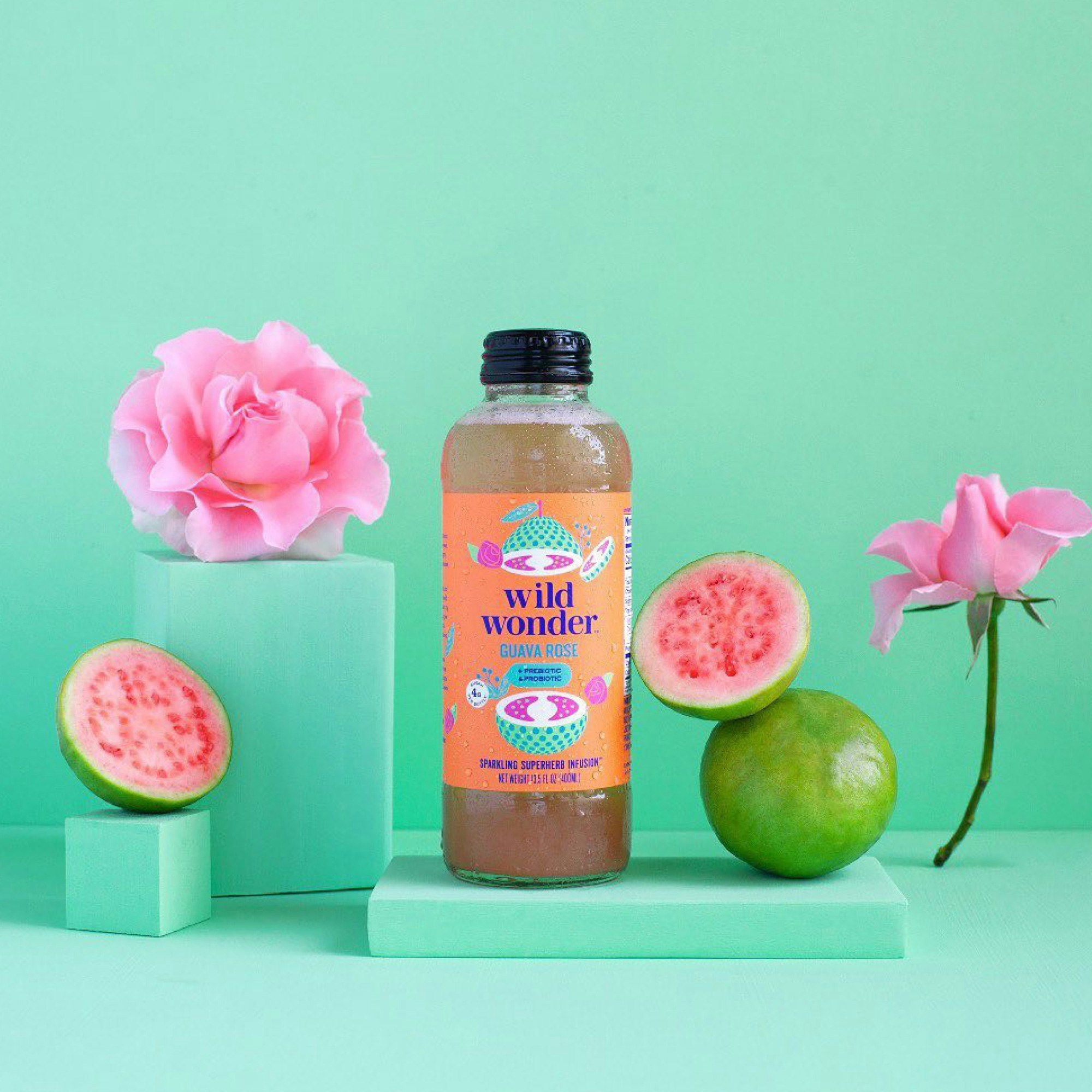 Bottle standing on a mint green seamless with pink flowers and pink guava fruit slices next to it.