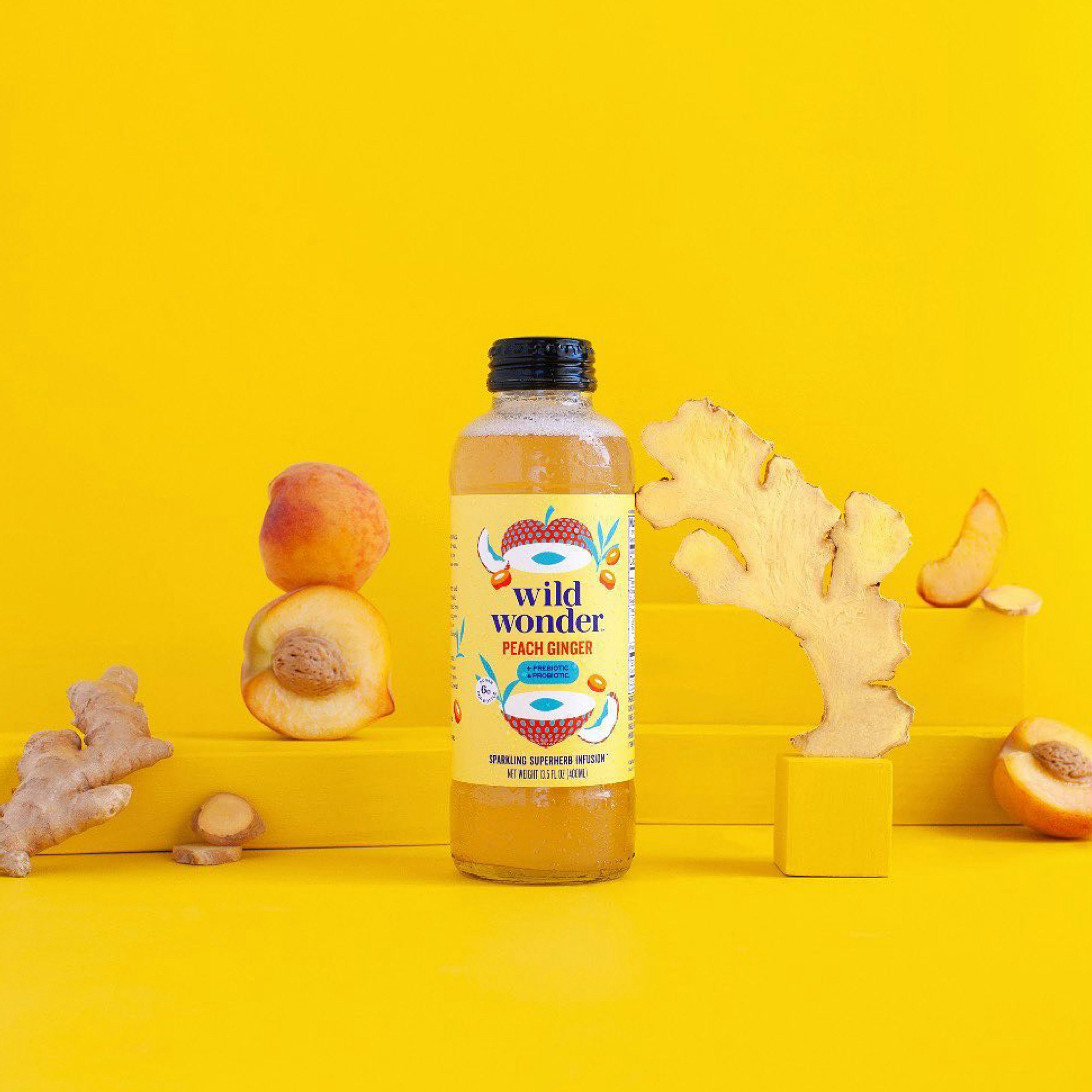 A glass bottle with a yellow beverage is standing on a yellow seamless surrounded by peaches and ginger.