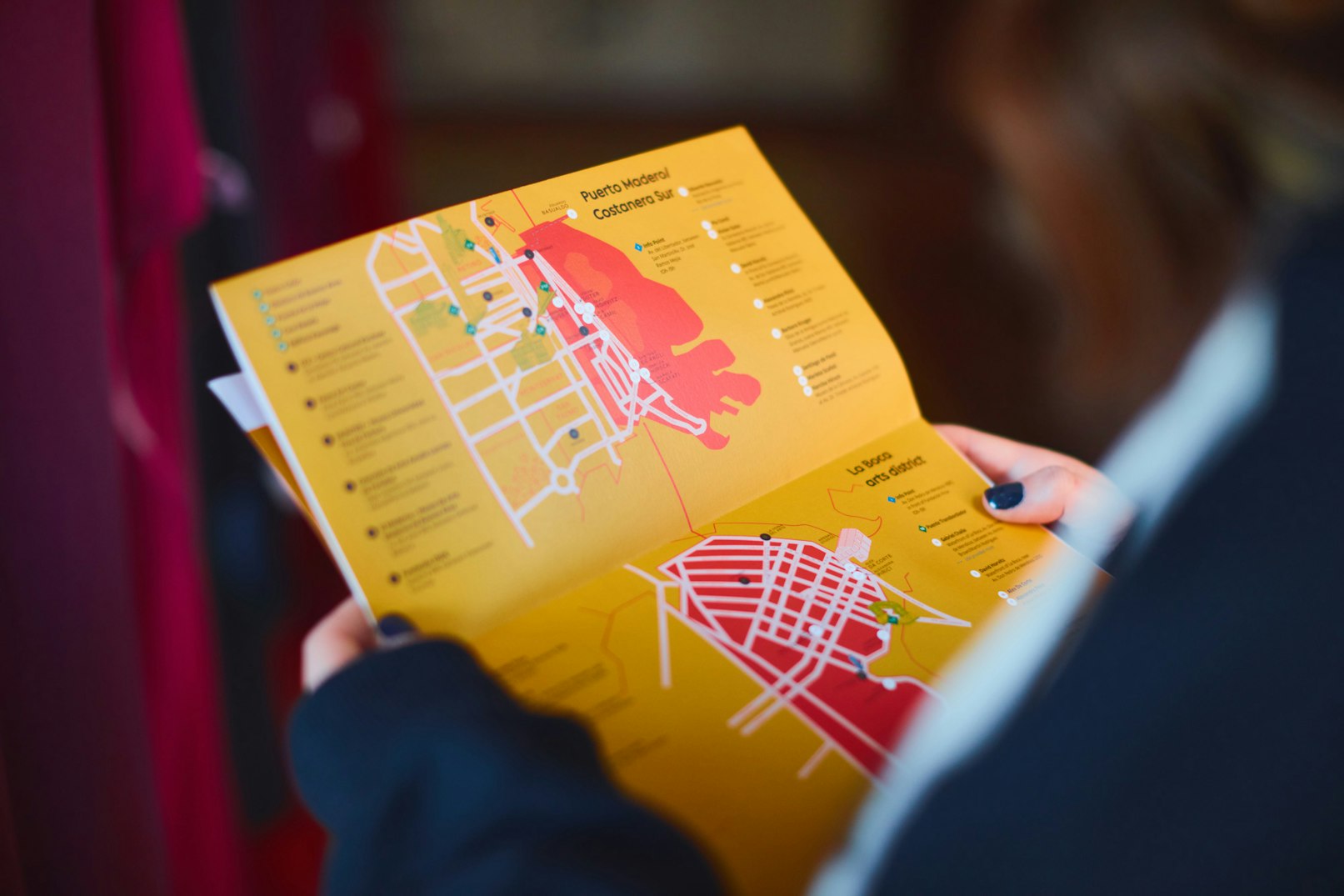 Image showing the close up of a person holding the Art Basel catalogue which is mostly dusty yellow and maps in red, white, dark blue.