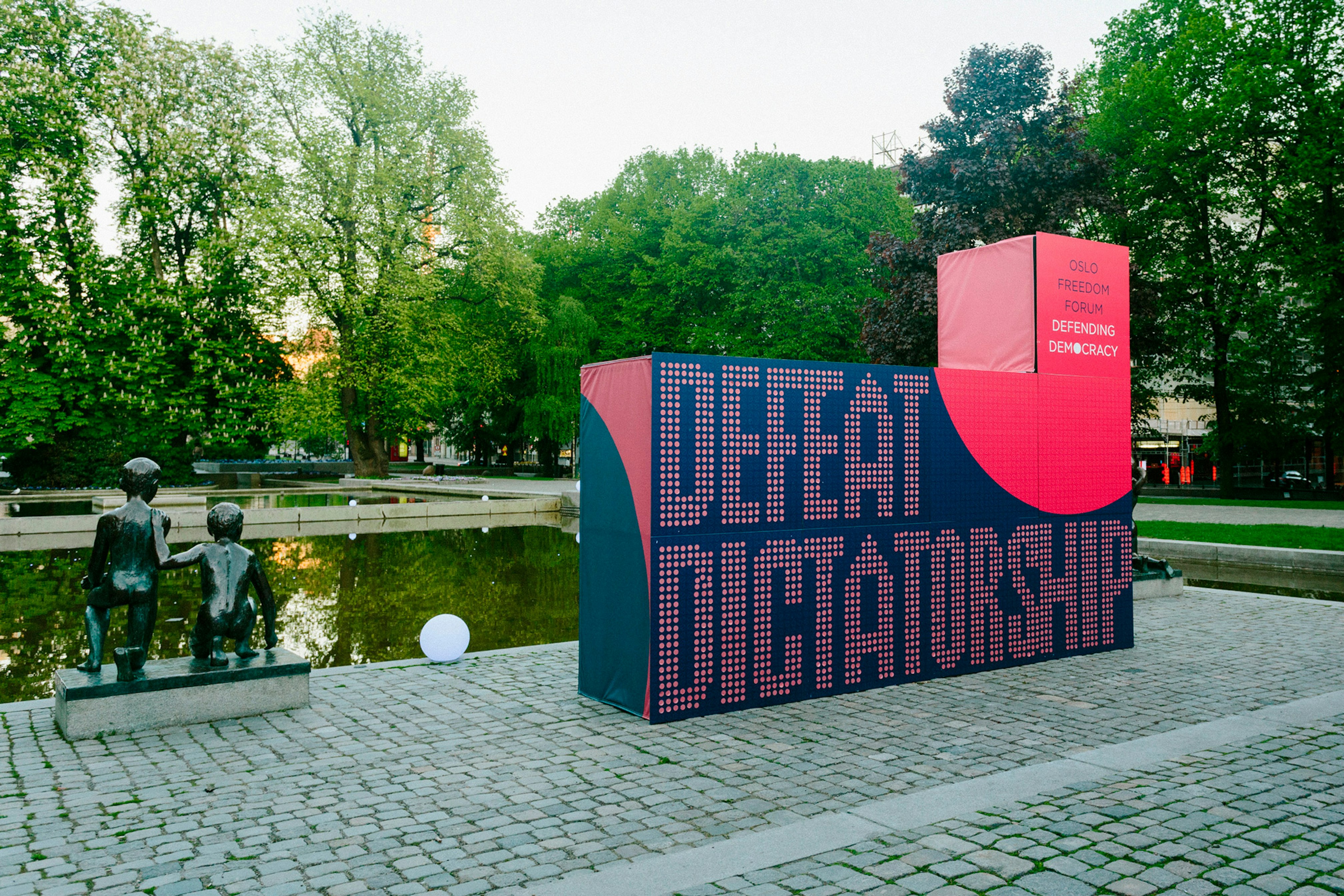 Image shows an L-shaped installation that is navy blue and coral-red. It says DEFEAT DICTATORSHIP on it. It's sits on a stone surface next to a statue of human figures and a pond. There are trees in the background. 