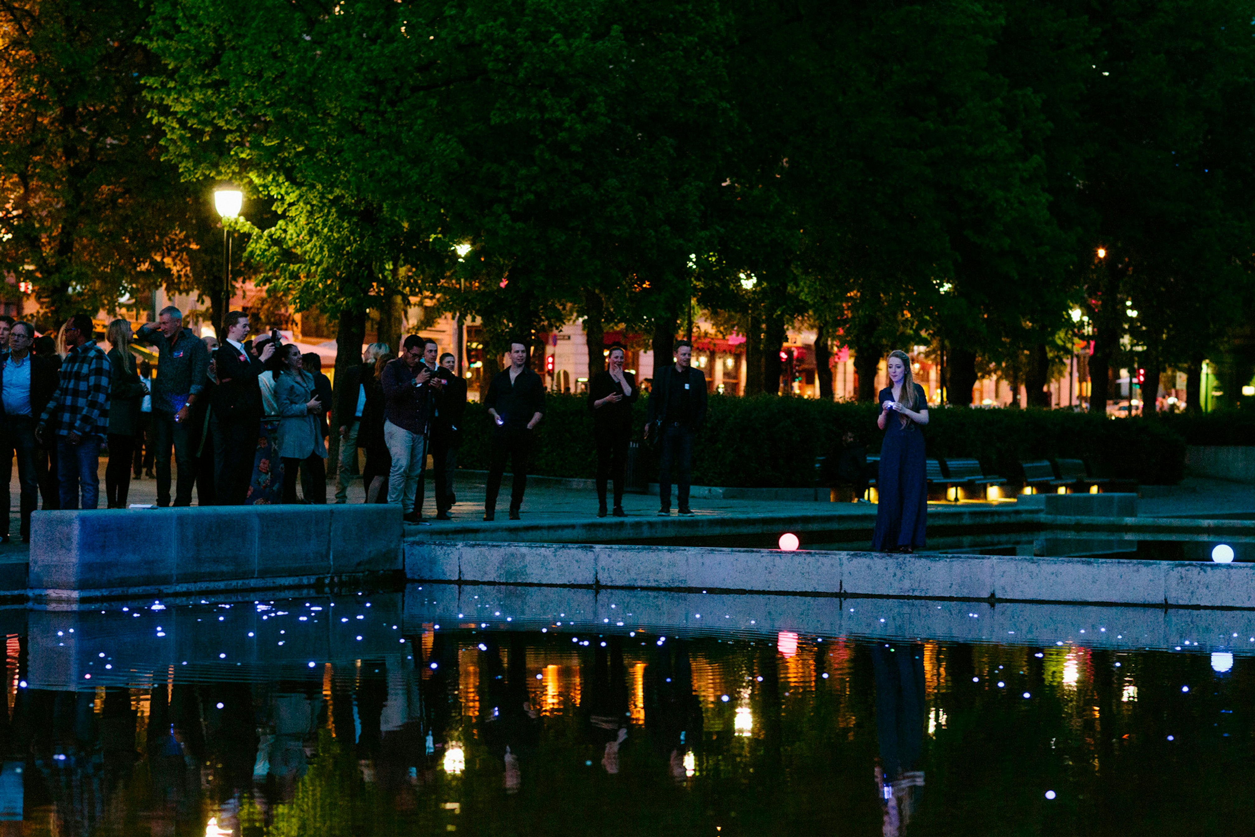 Image shows people in the dark surrounding a pond. There are many small illuminated blue balls in the pond. 