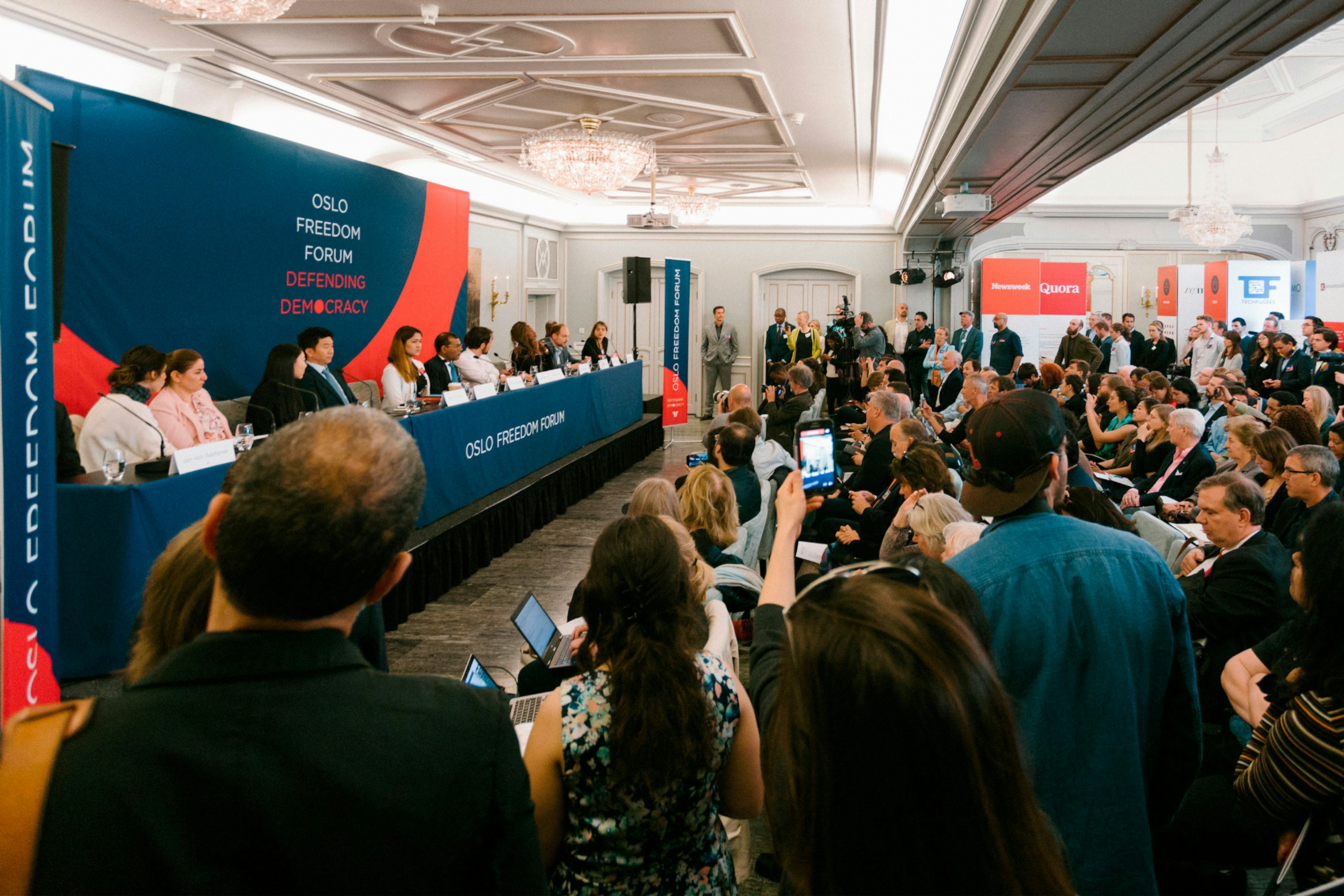 Image shows people crowded into a room with a panel sitting at a table in front of a blue and coral banner reading OSLO FREEDOM FORUM DEFENDING DEMOCRACY.