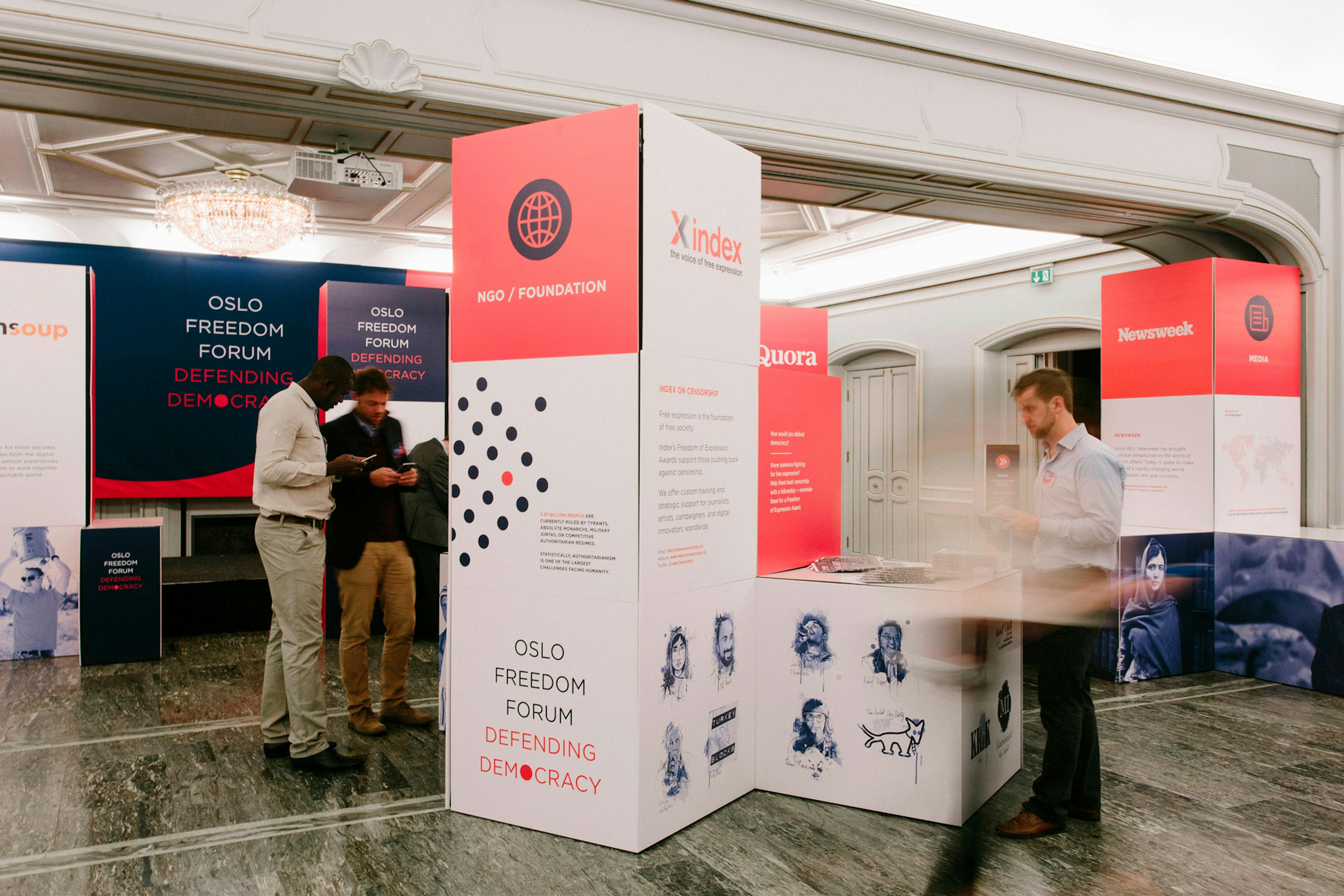 Image shows a 3D installation covered in coral, blue, and white graphics. Three people stand in the room around them. 