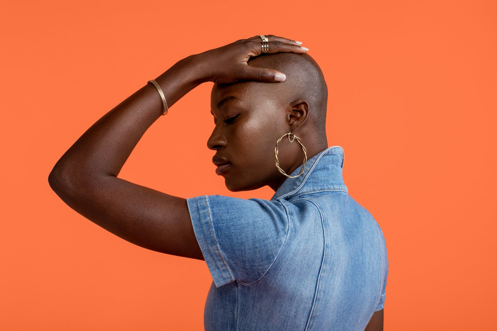 Image showing person's profile with their hand on their head showing off jewelry. They are wearing a blue denim jumpsuit and are standing against an orange seamless.