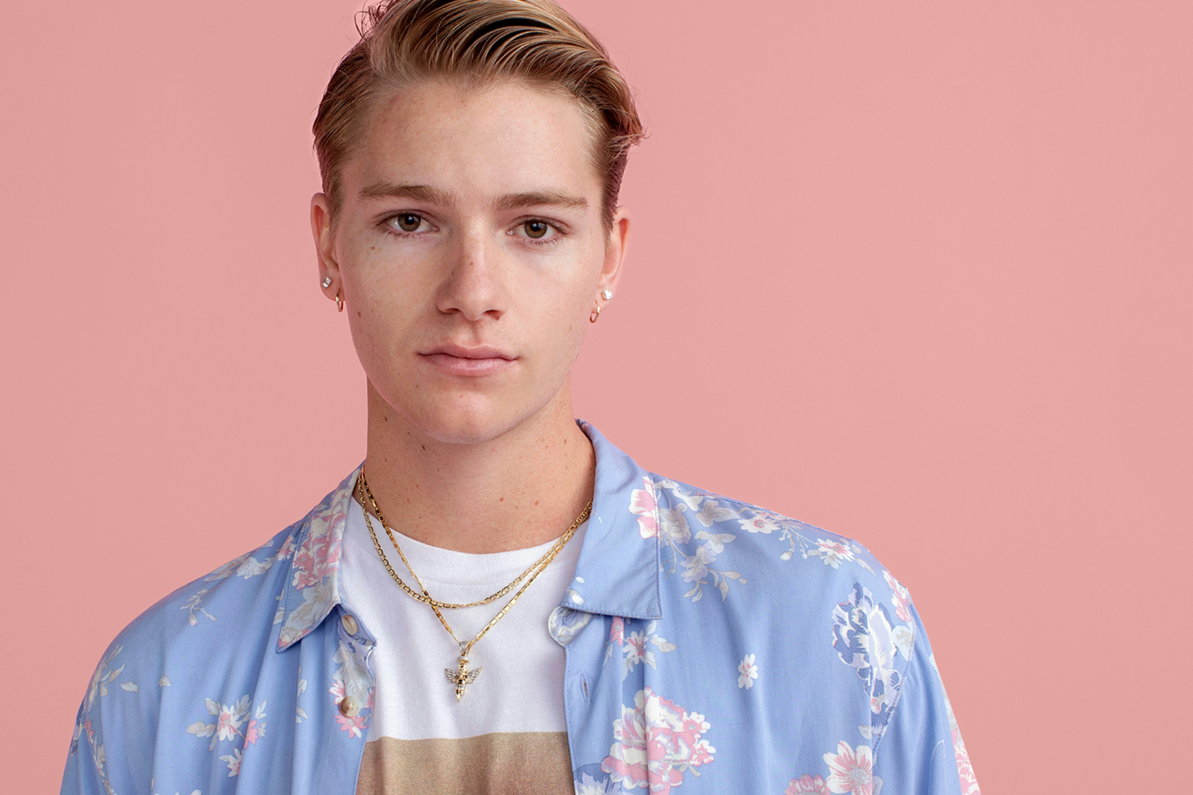 Image showing person on a soft pink seamless looking in camera. They are wearing a white teeshirt with an open shirt on top. The shirt is soft blue with a pink and blue floral pattern.