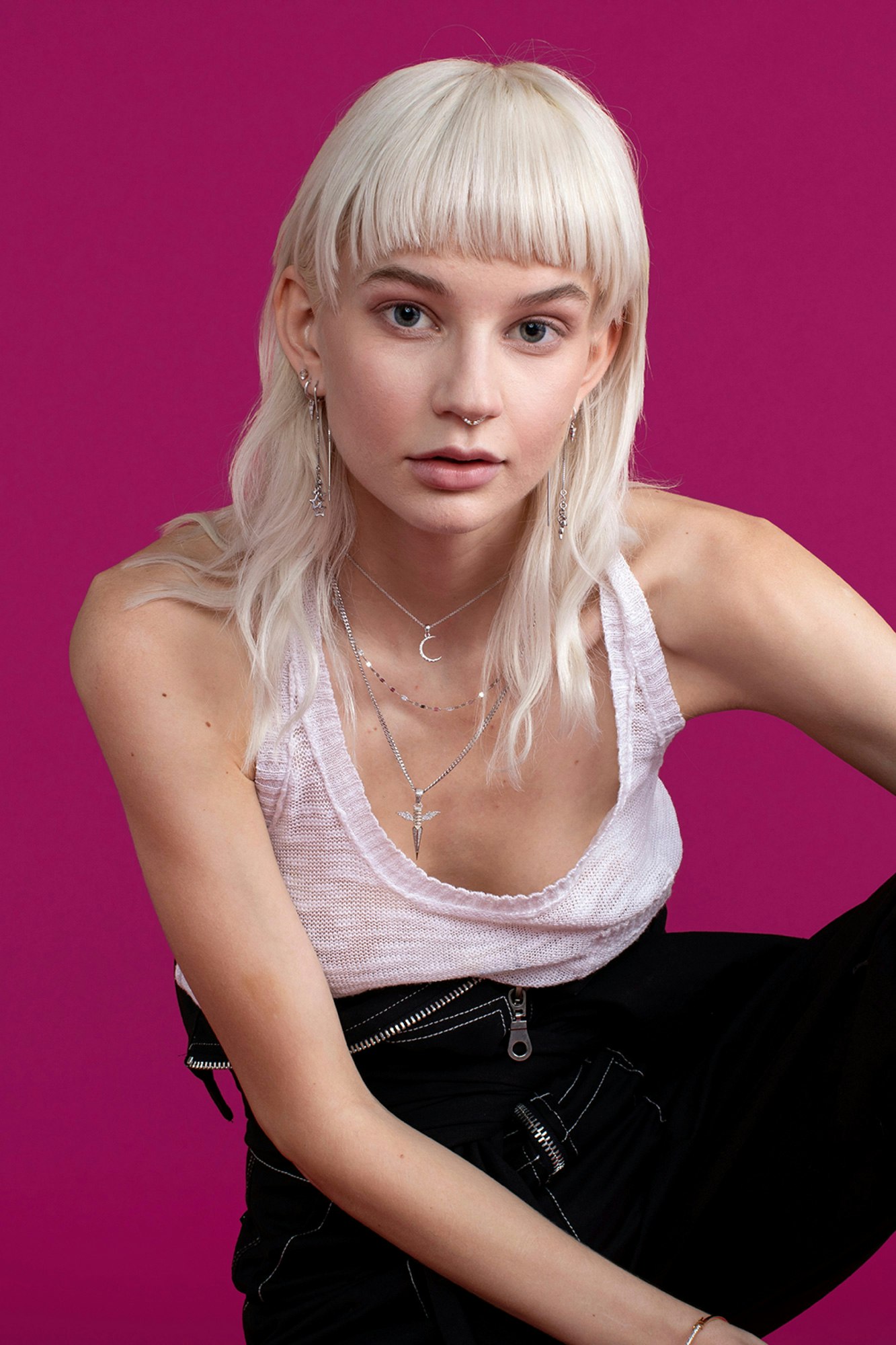 Image showing person against a berry colored seamless looking in camera. Their hair is nearly white and they are wearing a white tank-top and black jumpsuit tied around their waist.