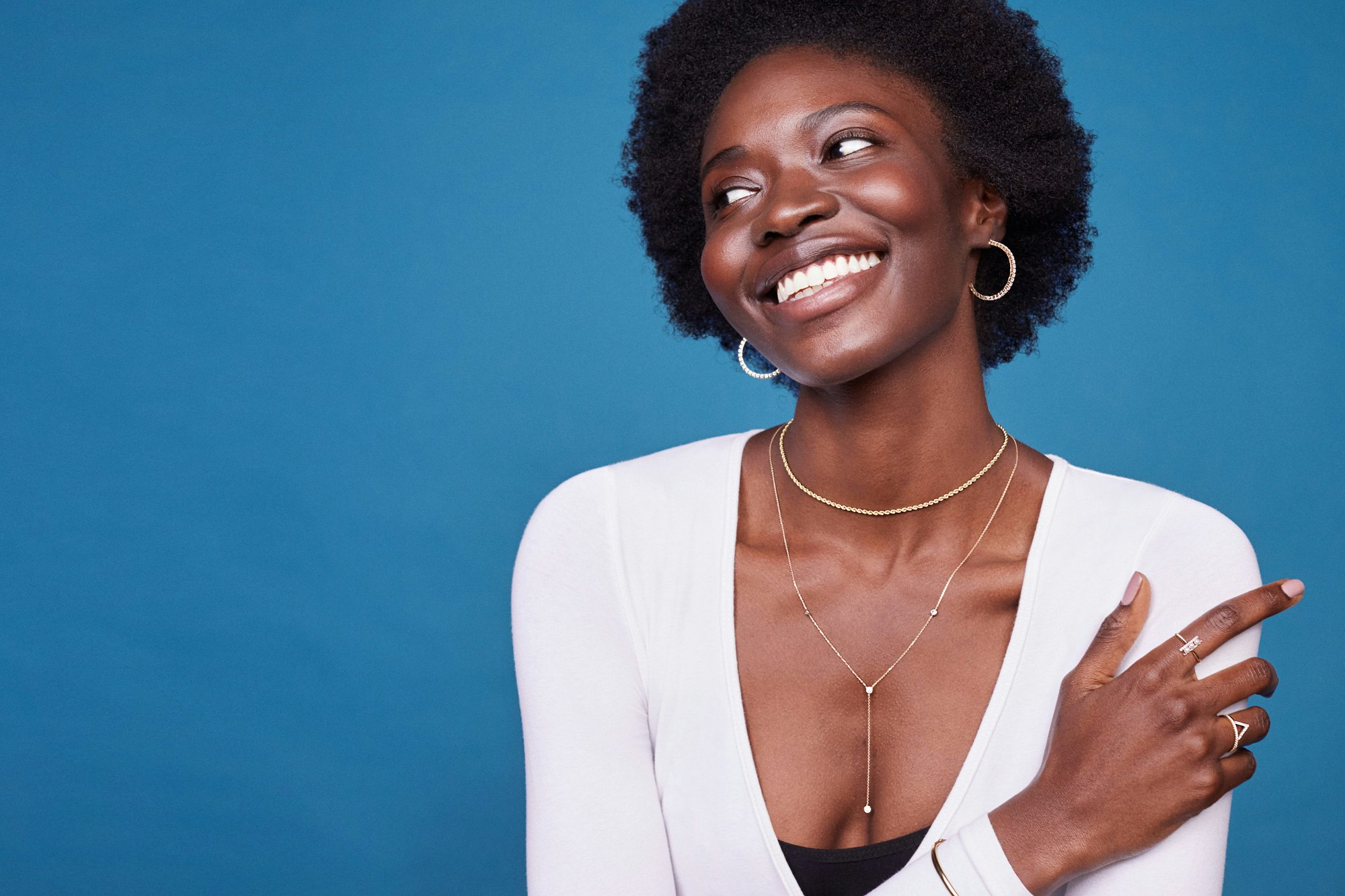 Image showing a person in a white wrap top standing against a dark blue seamless. They are looking to the side, smiling, looking happy.