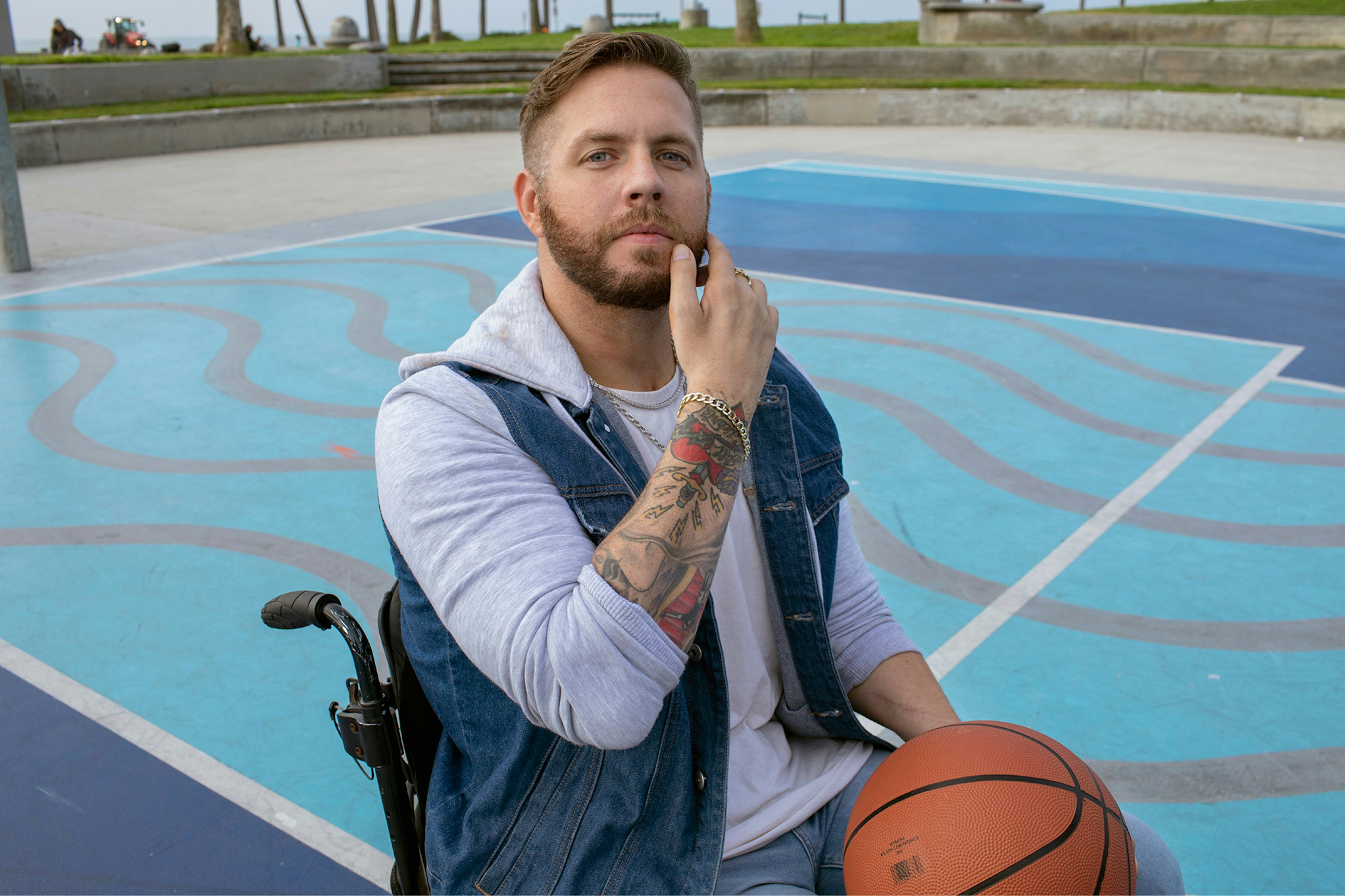 Image showing a person in a wheel chair on a basketball court. They are looking in camera, wearing a gray hoodie with a denim vest on top and holding an orange basket ball.