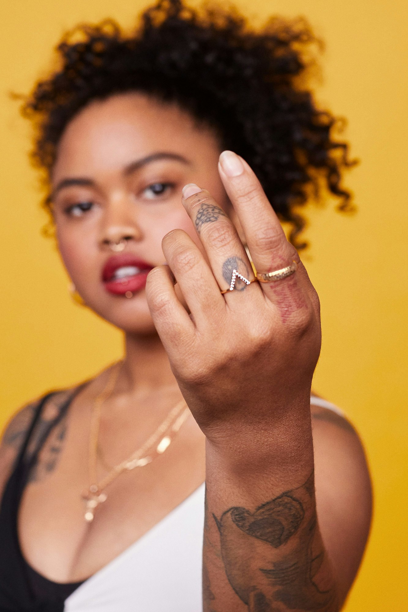 Image showing person in a black and white tank-top with their hand close to the camera showing tattoos on their fingers and arms. They are standing against a warm yellow seamless.