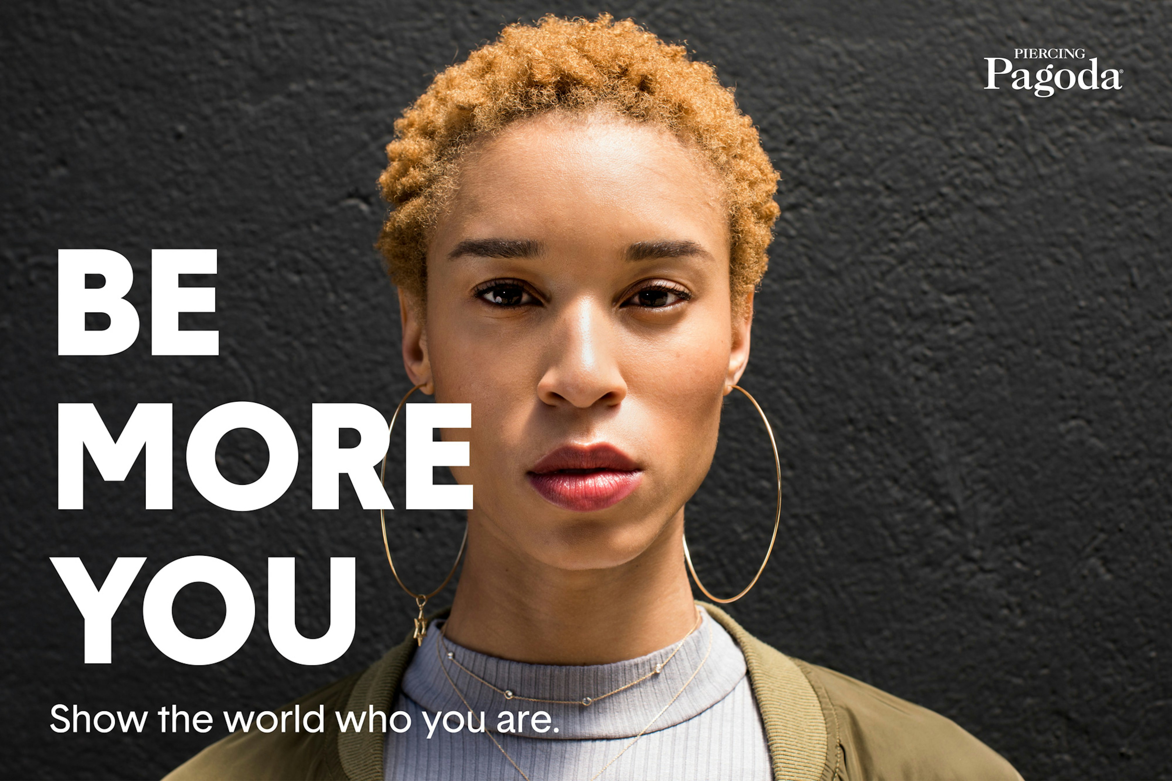 Image showing person standing against a dark gray wall, looking in camera. They have short hair and are wearing large gold hoop earrings, a gray mock turtleneck and an olive jacket. It says BE MORE YOU in white next to their face.