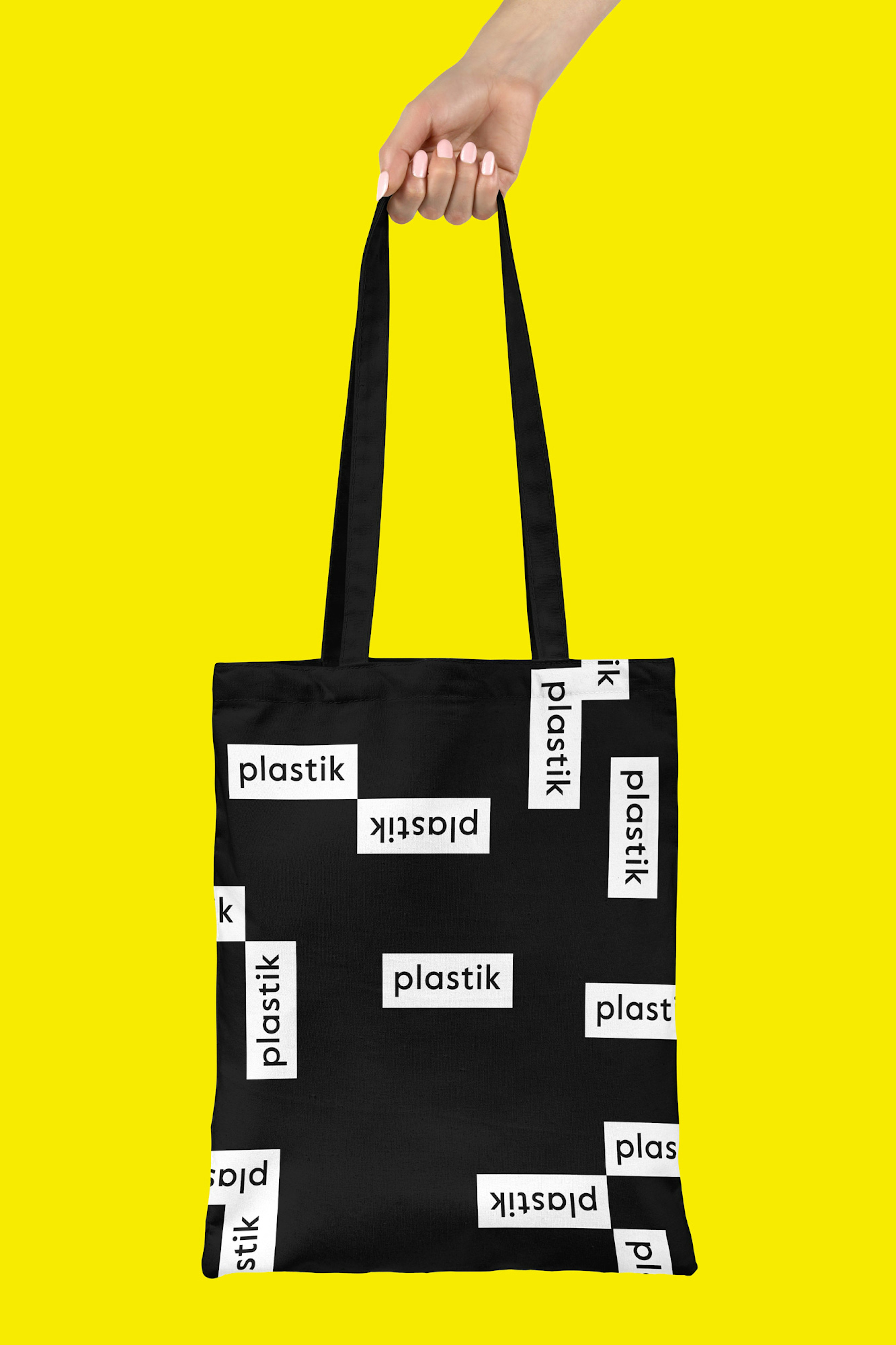 Image shows a hand holding a black tote bag covered in plastik logos in white rectangles. The background is bright yellow. 