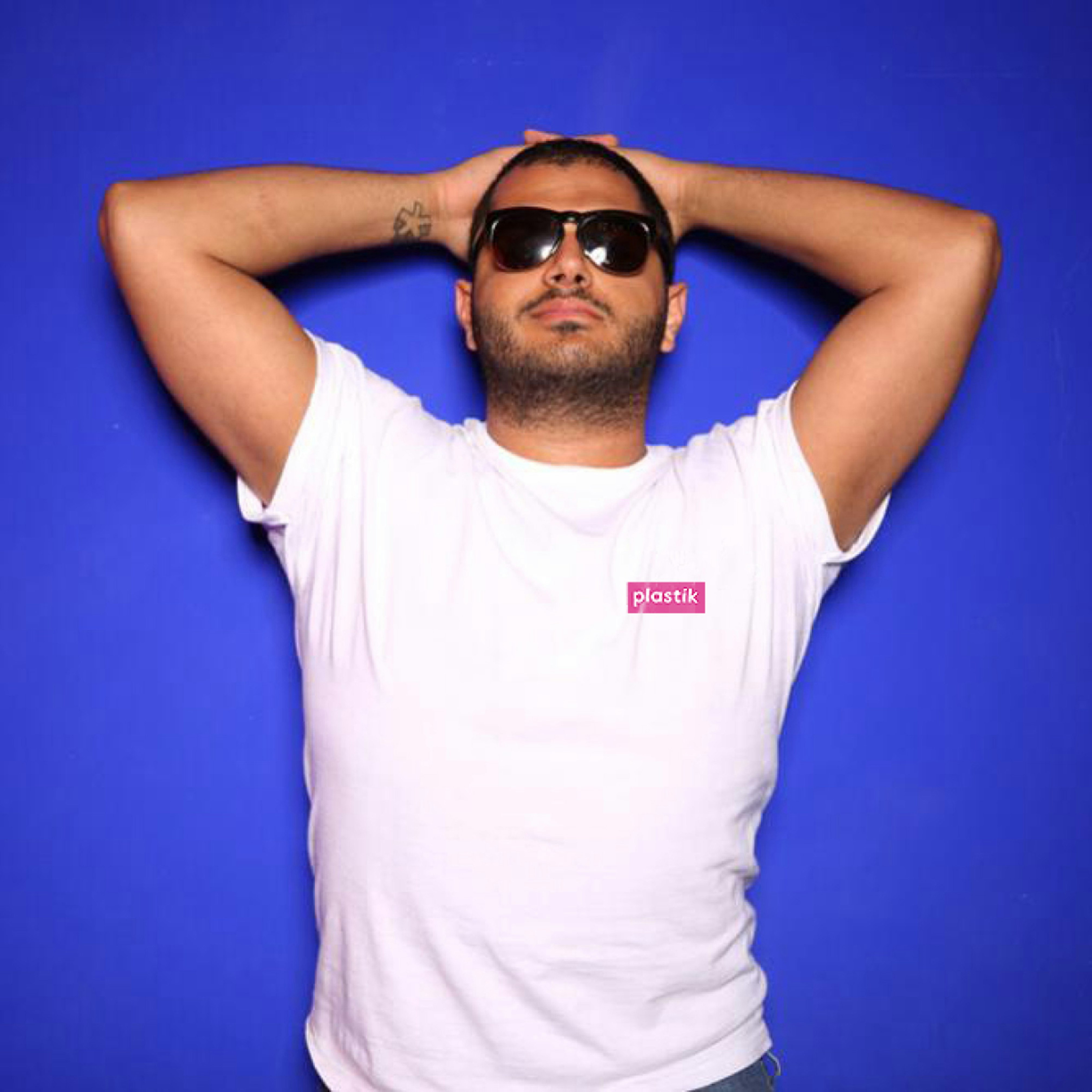 Image shows a person in black sunglasses with the arms clasped behind their head facing the camera on a bright blue background. They are wearing a white, short-sleeved tee shirt with a small plastik logo in a pink box on it. 