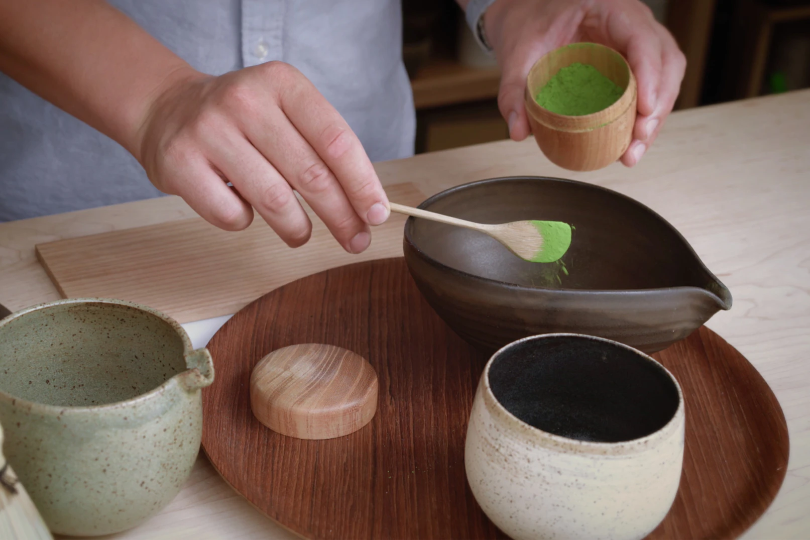 Image showing a pair of hands preparing a matcha using a wooden spoon to scoop the bright green powder into a ceramic bowl.