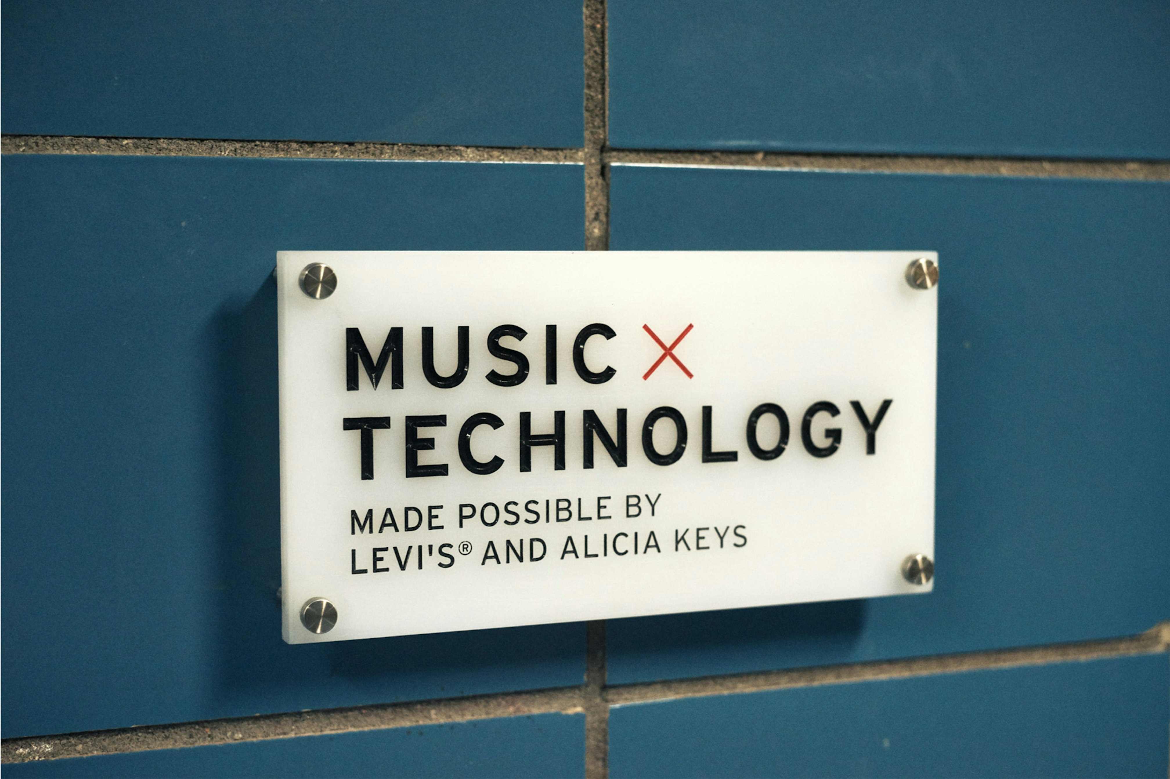 Image shows a close-up of a small white sign that says MUSIC X TECHNOLOGY MADE POSSIBLE BY LEVI'S AND ALICIA KEYS. It's mounted to a blue tile surface. 