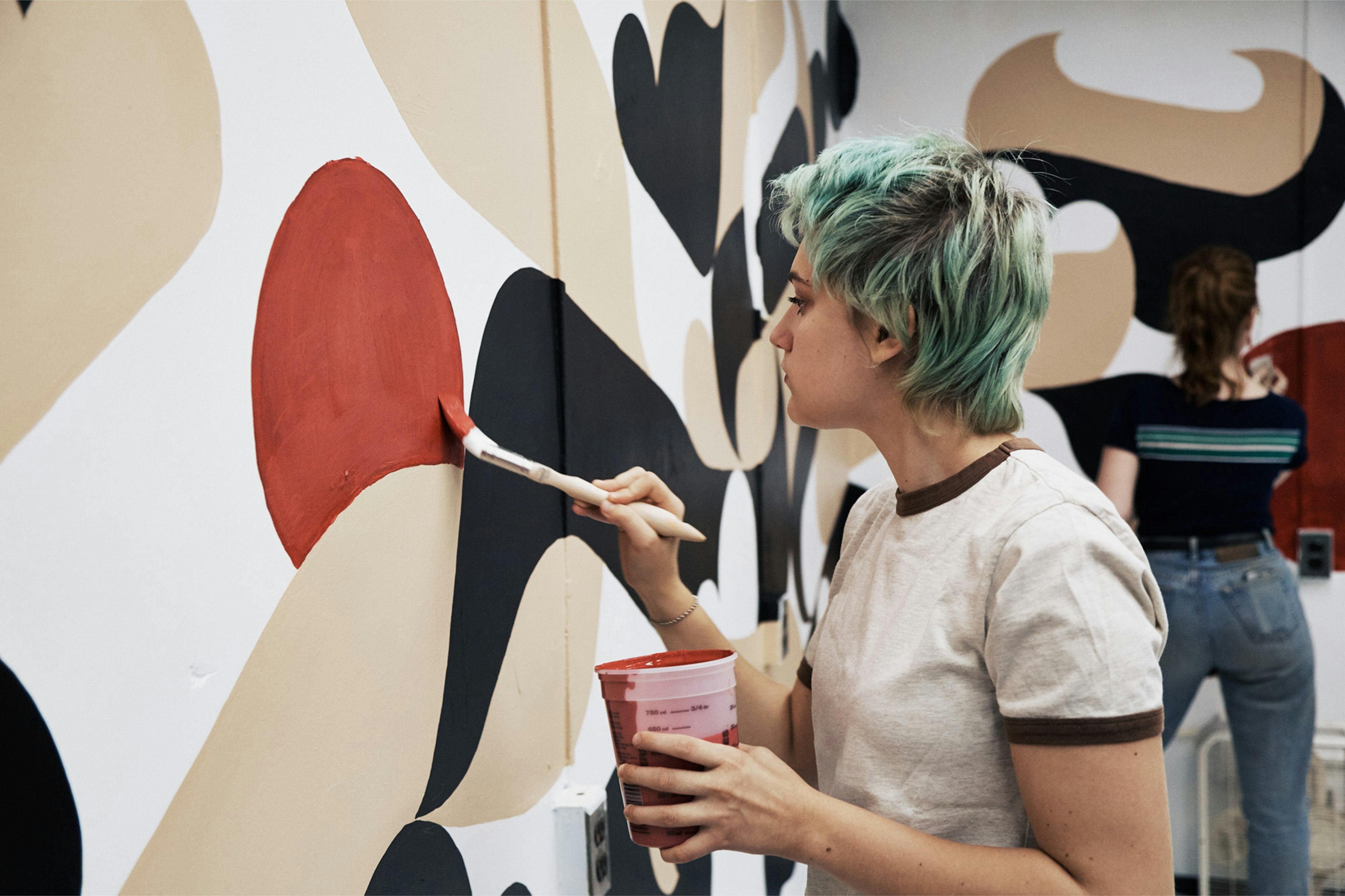 Image shows a person with short blue-green hair painting a mural on a white wall. They are working on a rust-colored shape. 