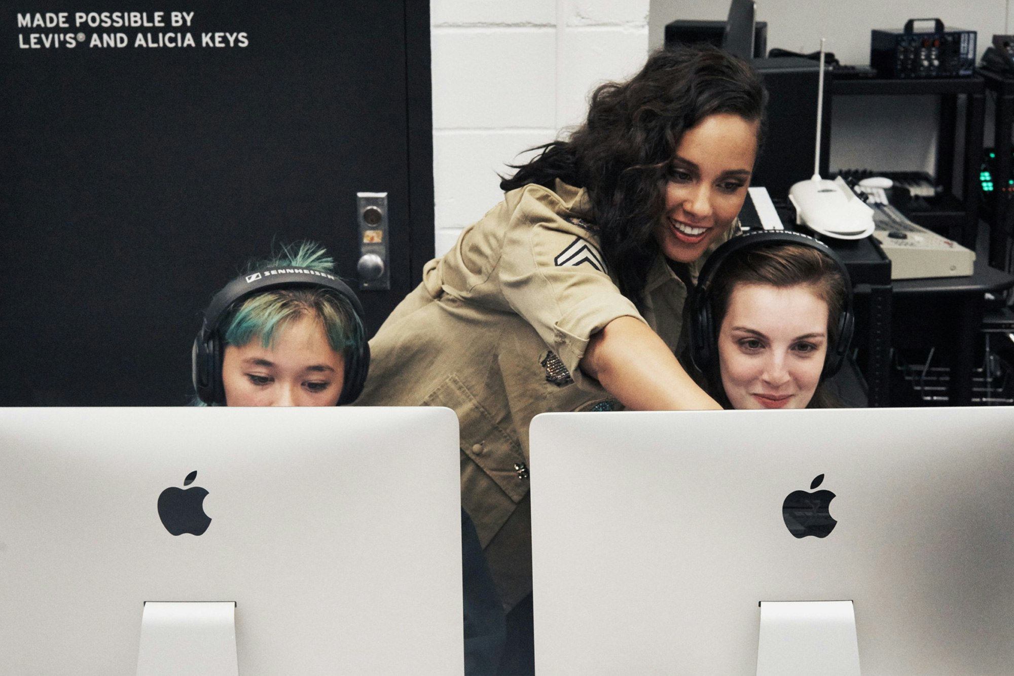 Image shows the back of two desktop Macs in the foreground. Behind them are two young people looking at the computer screens. Alicia Keys is behind them, pointing at something on the righthand computer screen. In the upper left corner of the image is text that reads MADE POSSIBLE BY LEVI'S AND ALICIA KEYS.