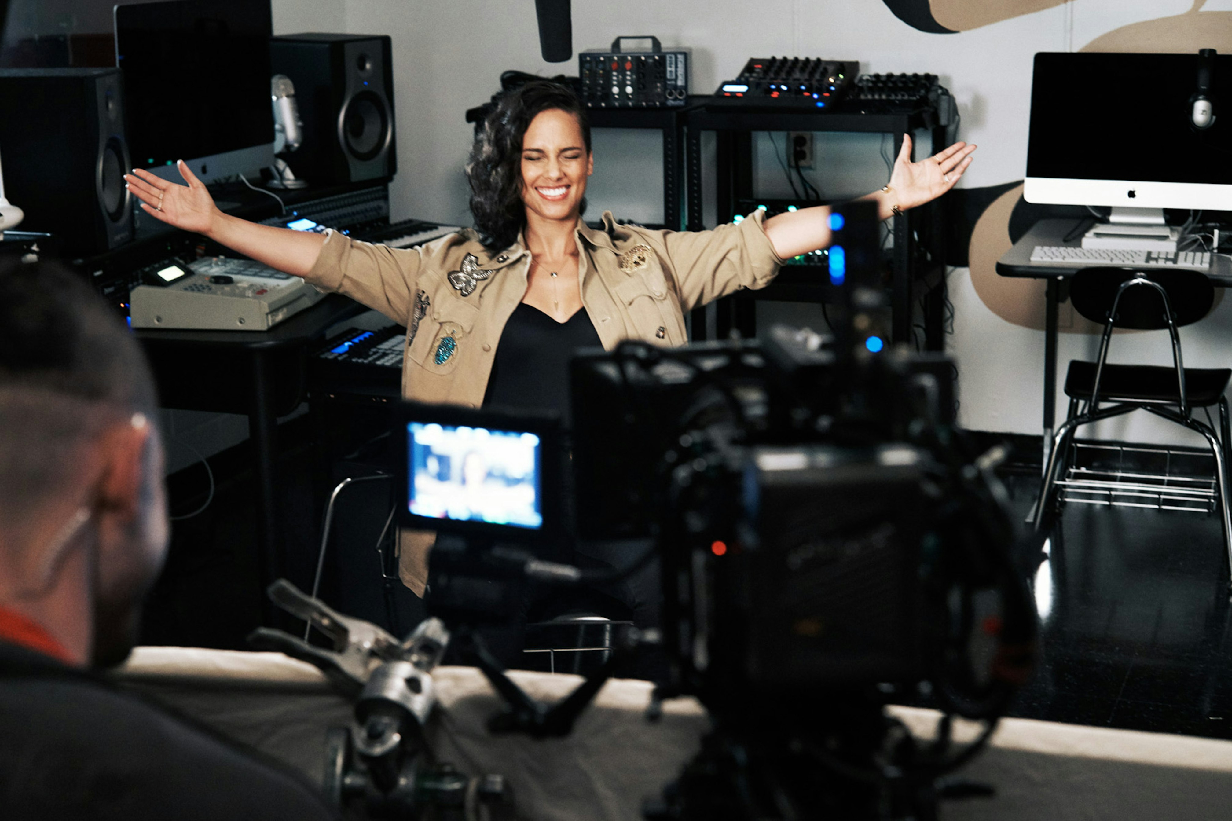 Image shows Alicia Keys with her eyes closed, smiling, and with her harms stretched out on either side of her. She's wearing a beige-colored jacket. 