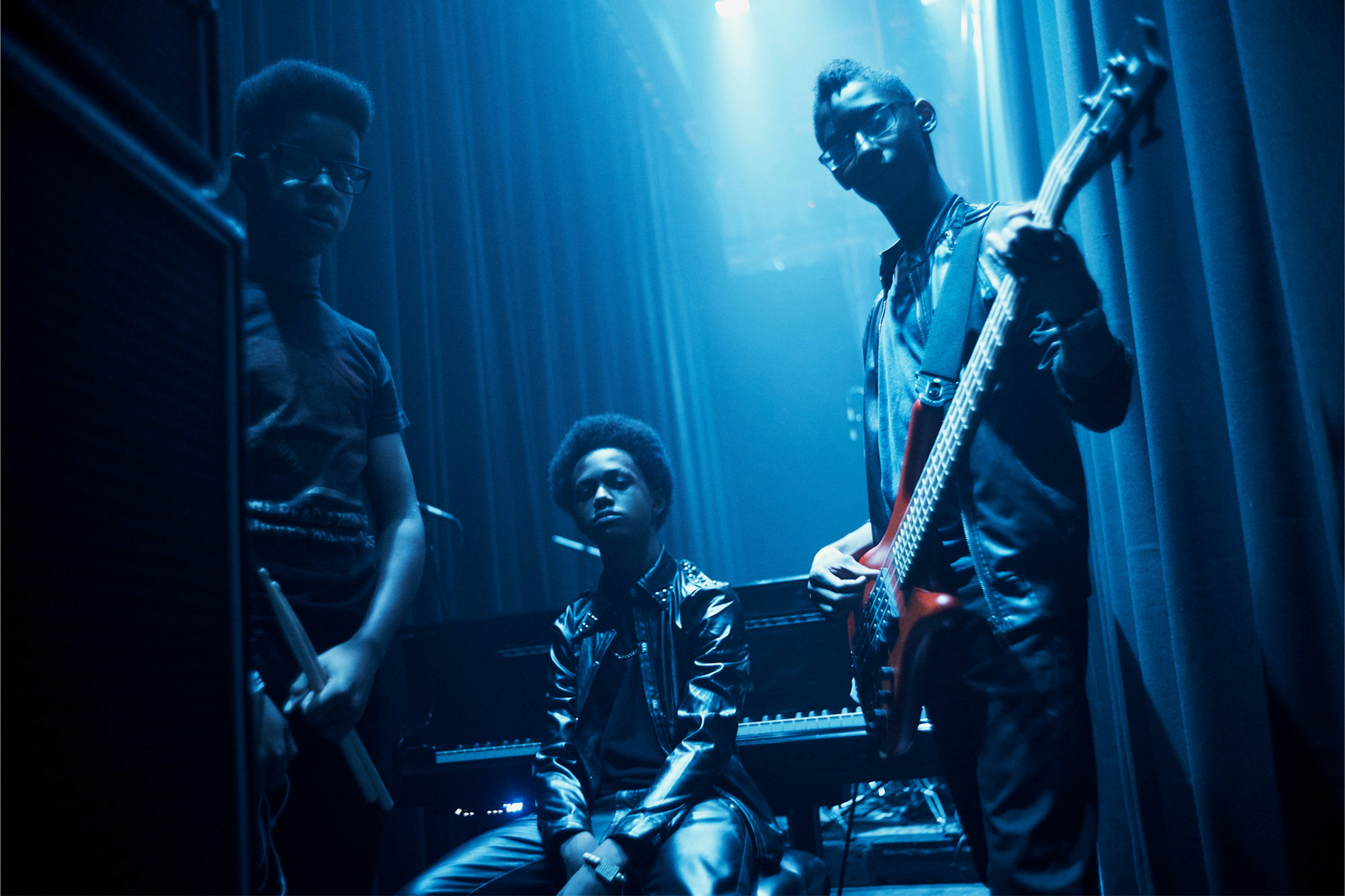Image shows three young people in a blue-lit space. One holds drumsticks, one holds a bass guitar, and the other is sitting in front of a keyboard. 