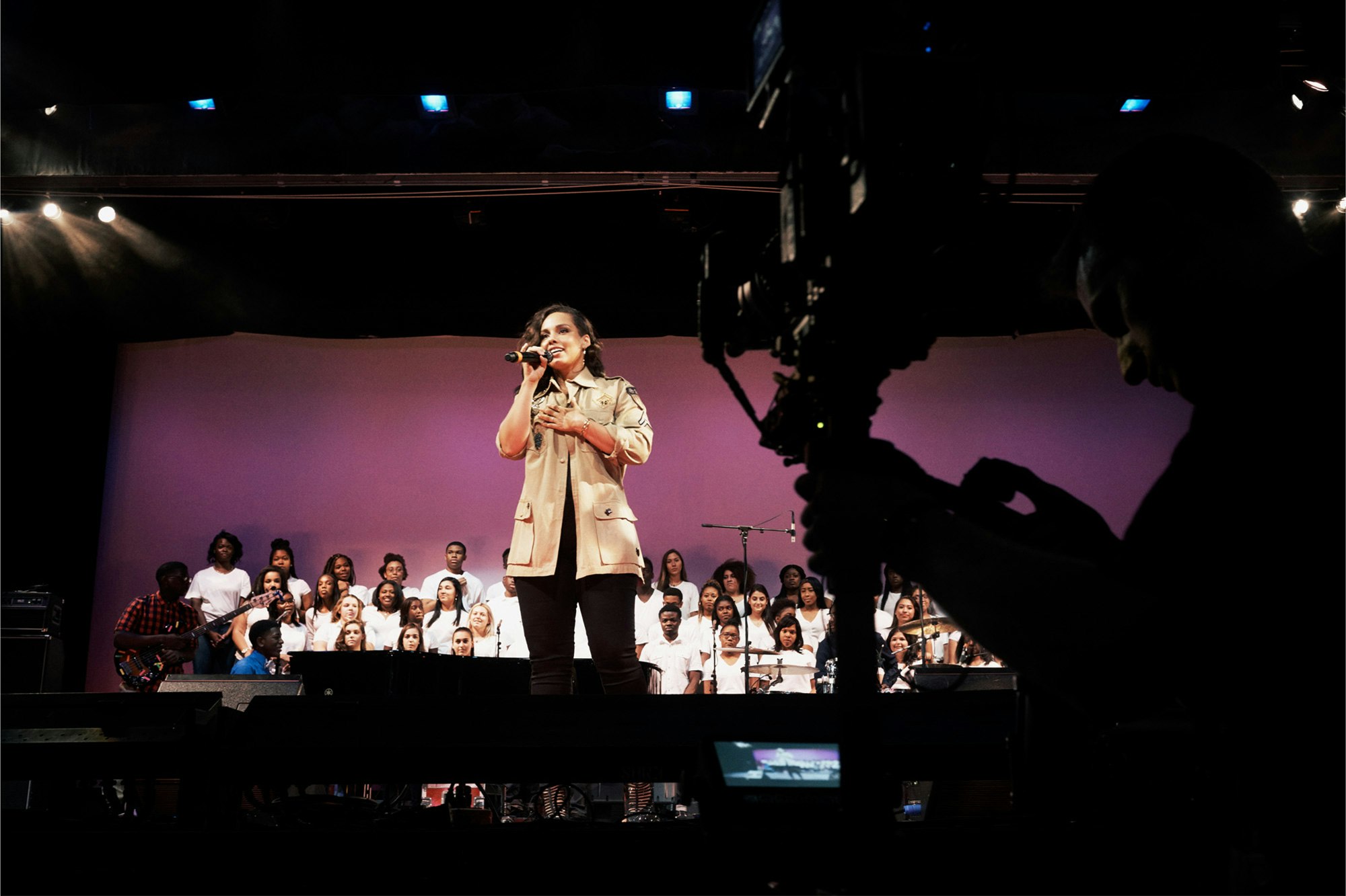 Image shows Alicia Keys on a stage holding a microphone to her face. She wears a beige jacket and pants.