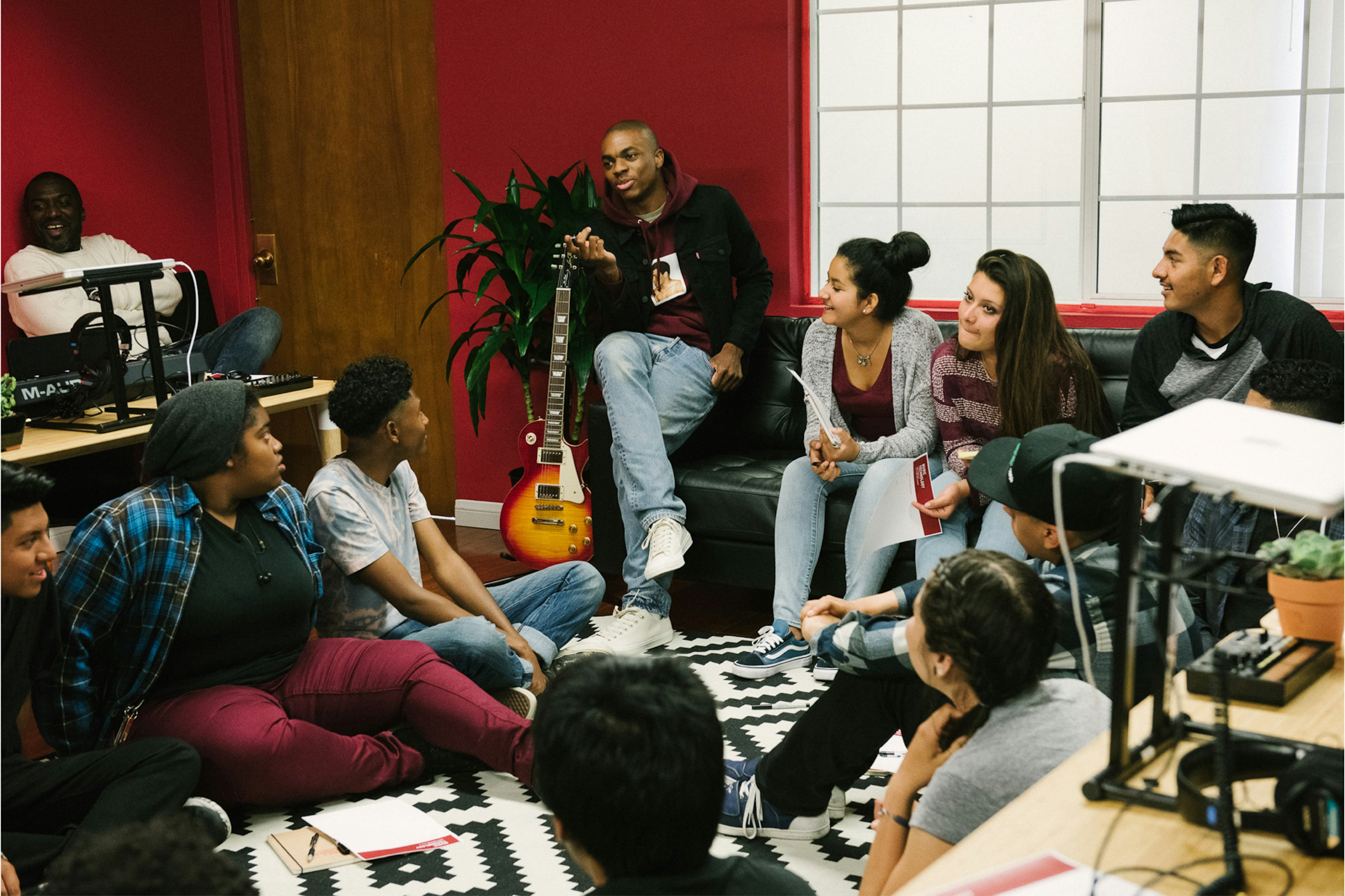 Image shows a group of young people sitting in a room with Vince Staples. Most of them are looking toward him, and he sits on the arm of a leather couch next to a guitar. There is a graphic black and white rug on the floor, where some of the young people are sitting. The walls in the room are red. 