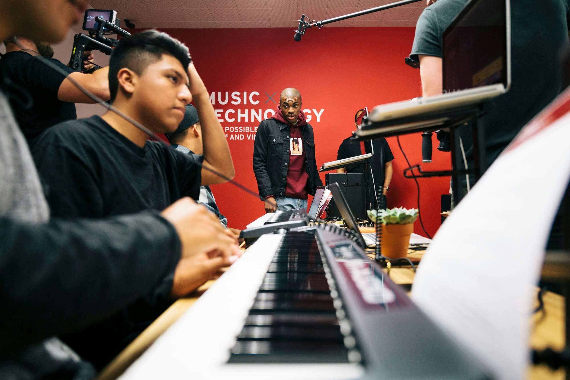 Image shows a keyboard (instrument) in the foreground and a young person working on a laptop. Vince is in the background, and behind him is a red wall that reads MUSIC x TECHNOLOGY.