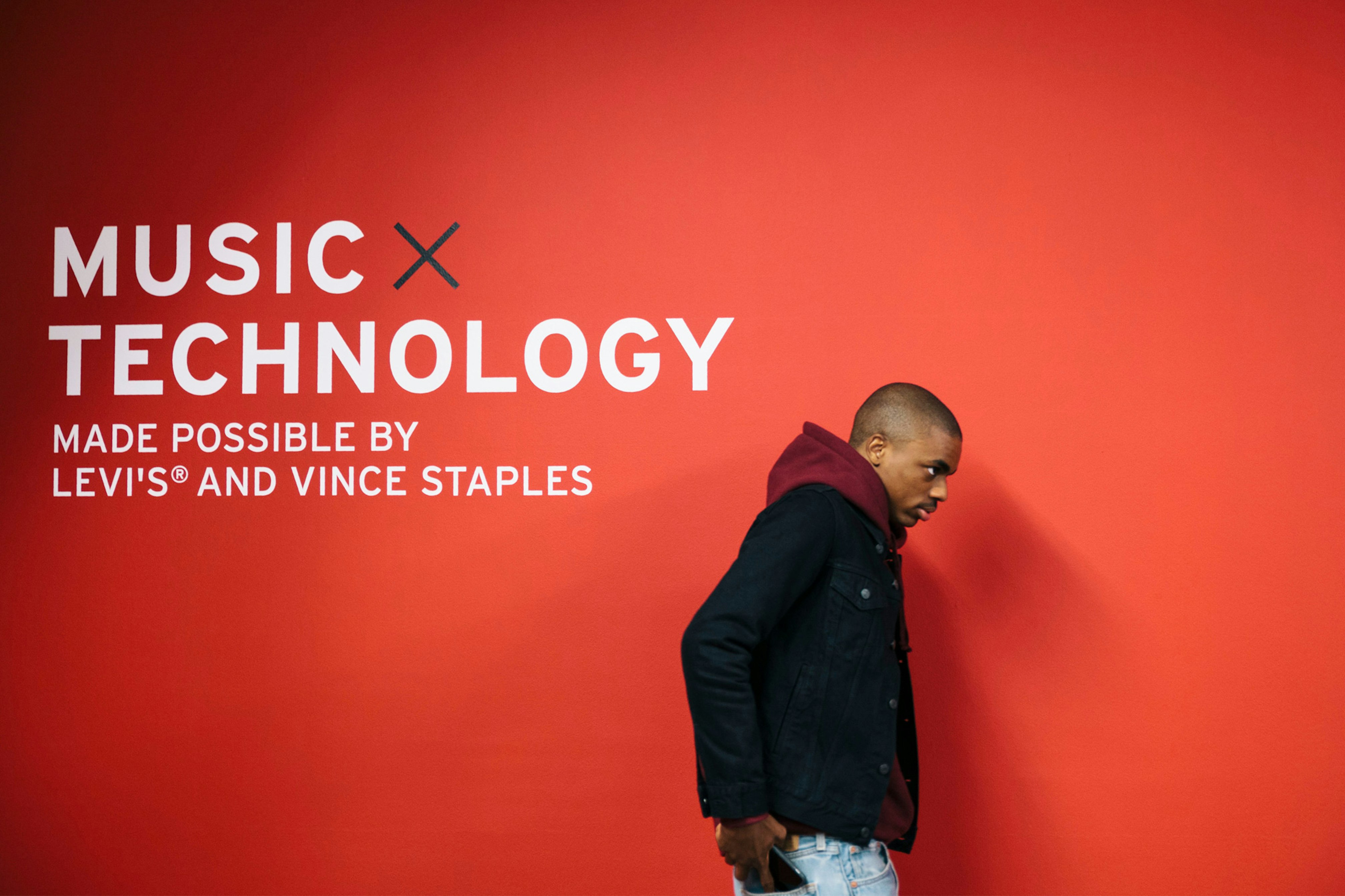 Image shows Vince Staples walking, facing the righthand side of the photograph. Behind him is a bright red wall that says MUSIC x TECHNOLOGY MADE POSSIBLE BY LEVI'S AND VINCE STAPLES in white sans serif text.  