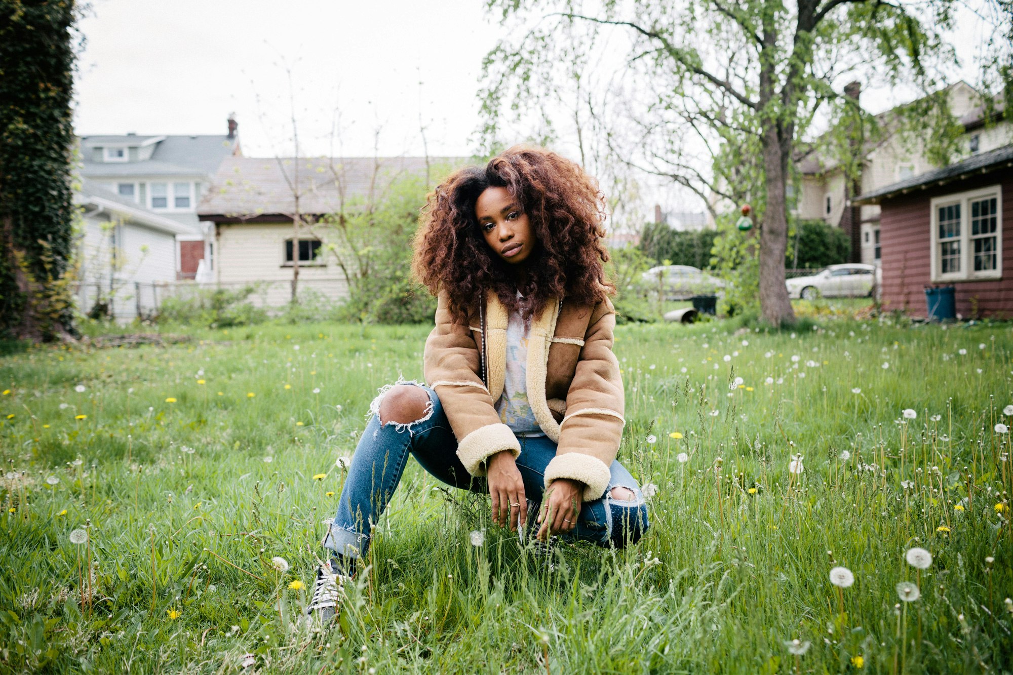 Image shows a portrait of SZA crouching in green grass. She wears a tan sherpa jacket with blue jeans ripped at the knees. She looks straight into the camera. Her curly brown hair is worn down.