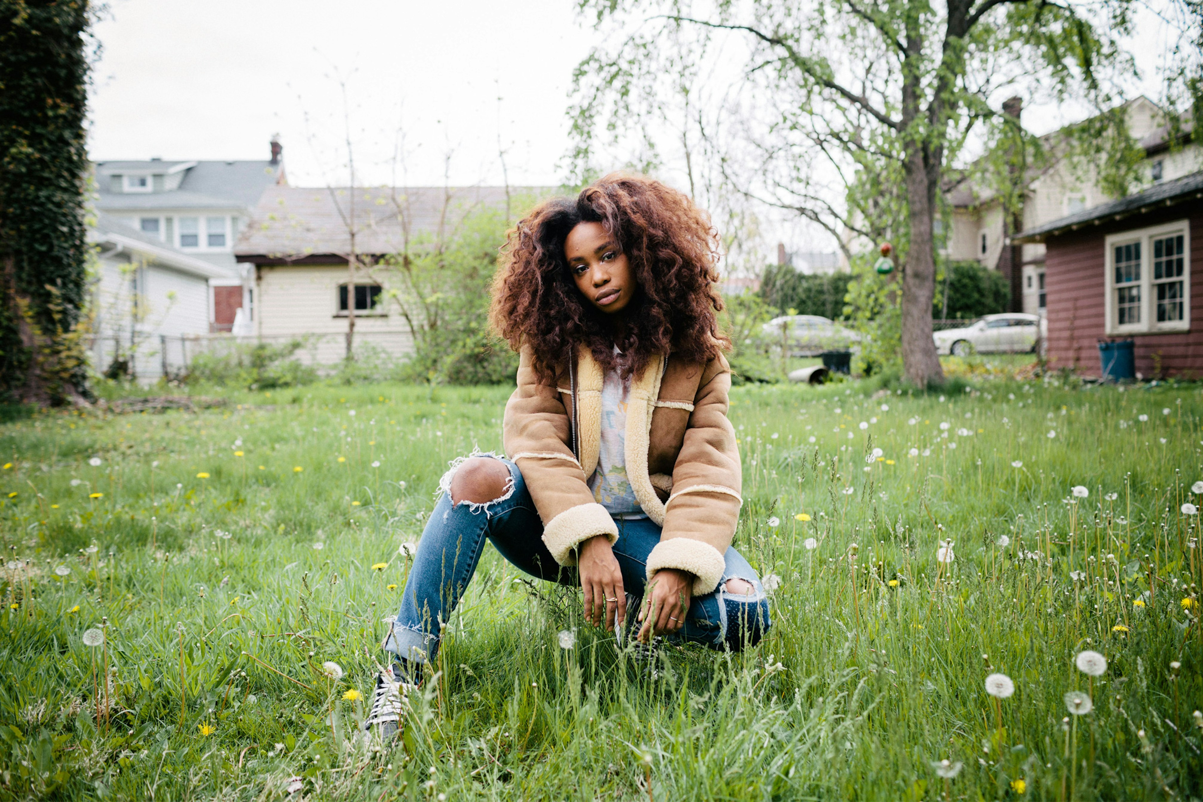 Image shows a portrait of SZA crouching in green grass. She wears a tan sherpa jacket with blue jeans ripped at the knees. She looks straight into the camera. Her curly brown hair is worn down. 