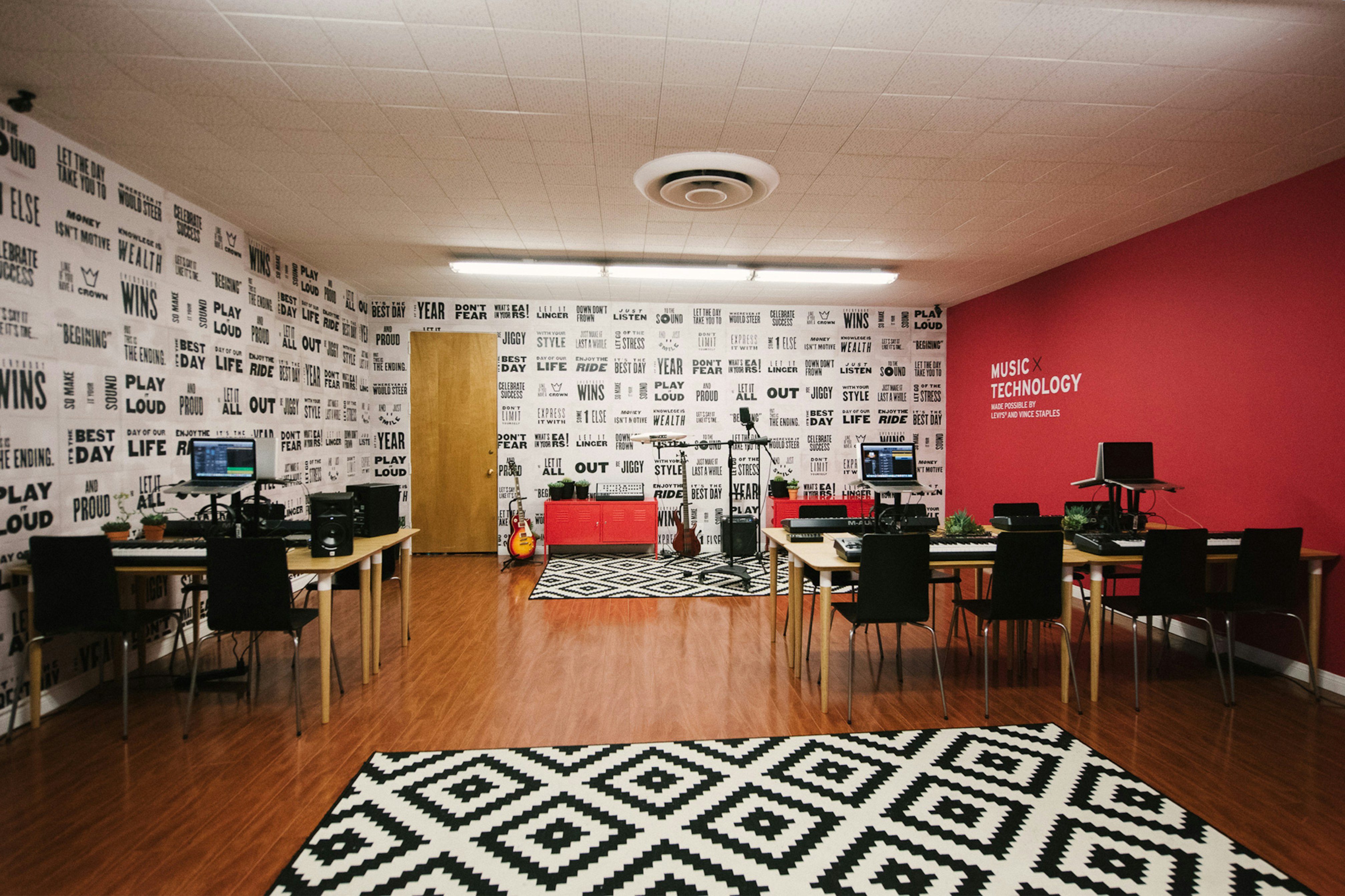 Image shows the interior of a room. The wall on the righthand side is red. There are desks and chairs and a door on the far wall. There is a graphic black and white rug on the floor in the foreground and black and white posters completed covering the left and back walls. 