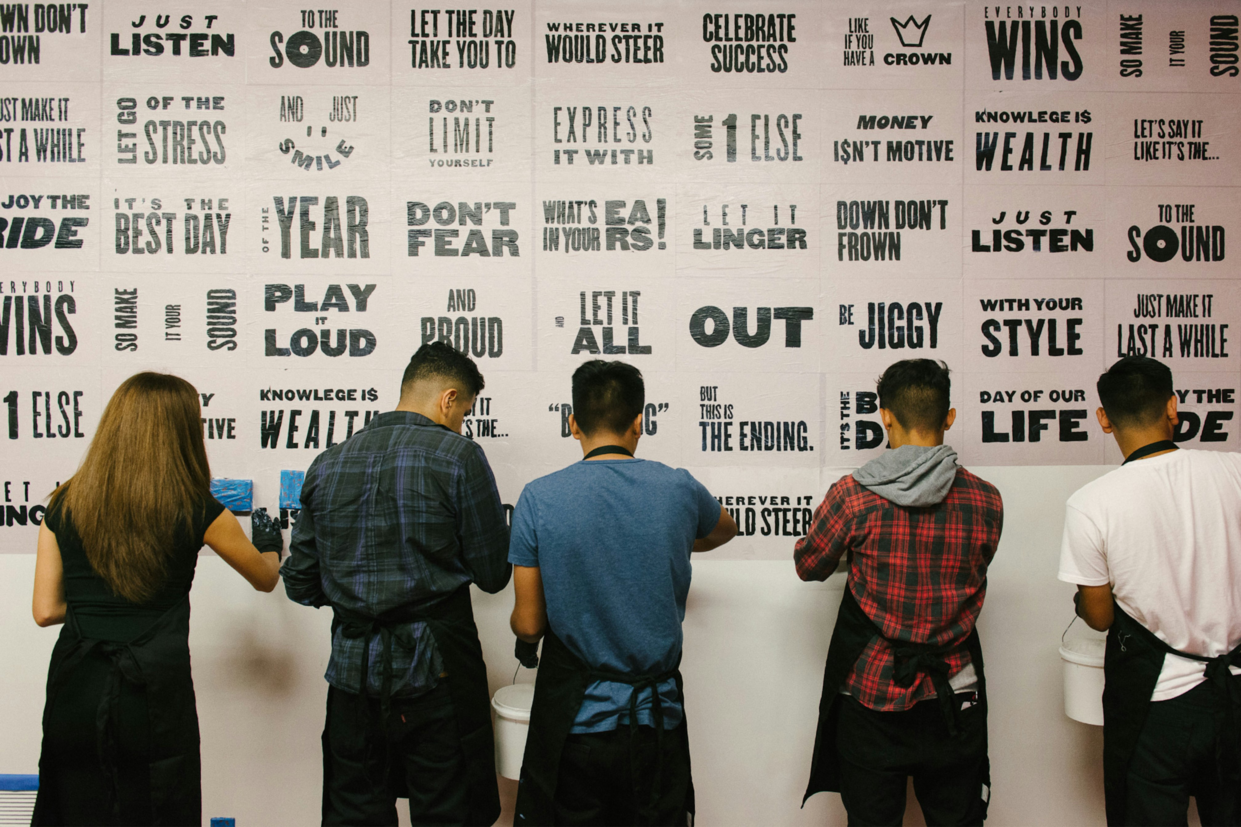 Image shows five people from behind working on wheatpasting the black and white text posters to the wall. 