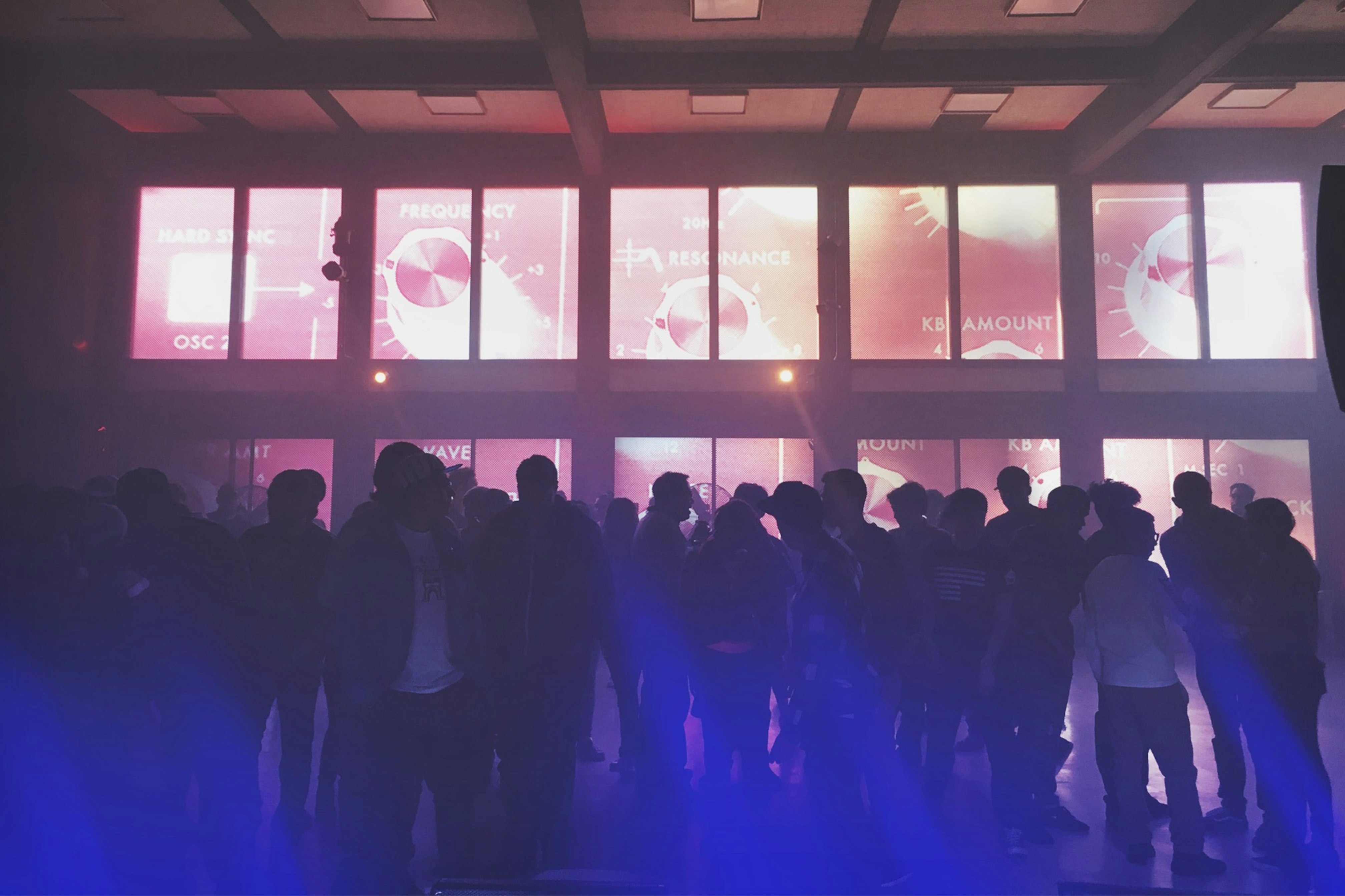 Image shows a silhouetted crowd standing in a room with large windows with graphic screens. The foreground glows bright blue. 