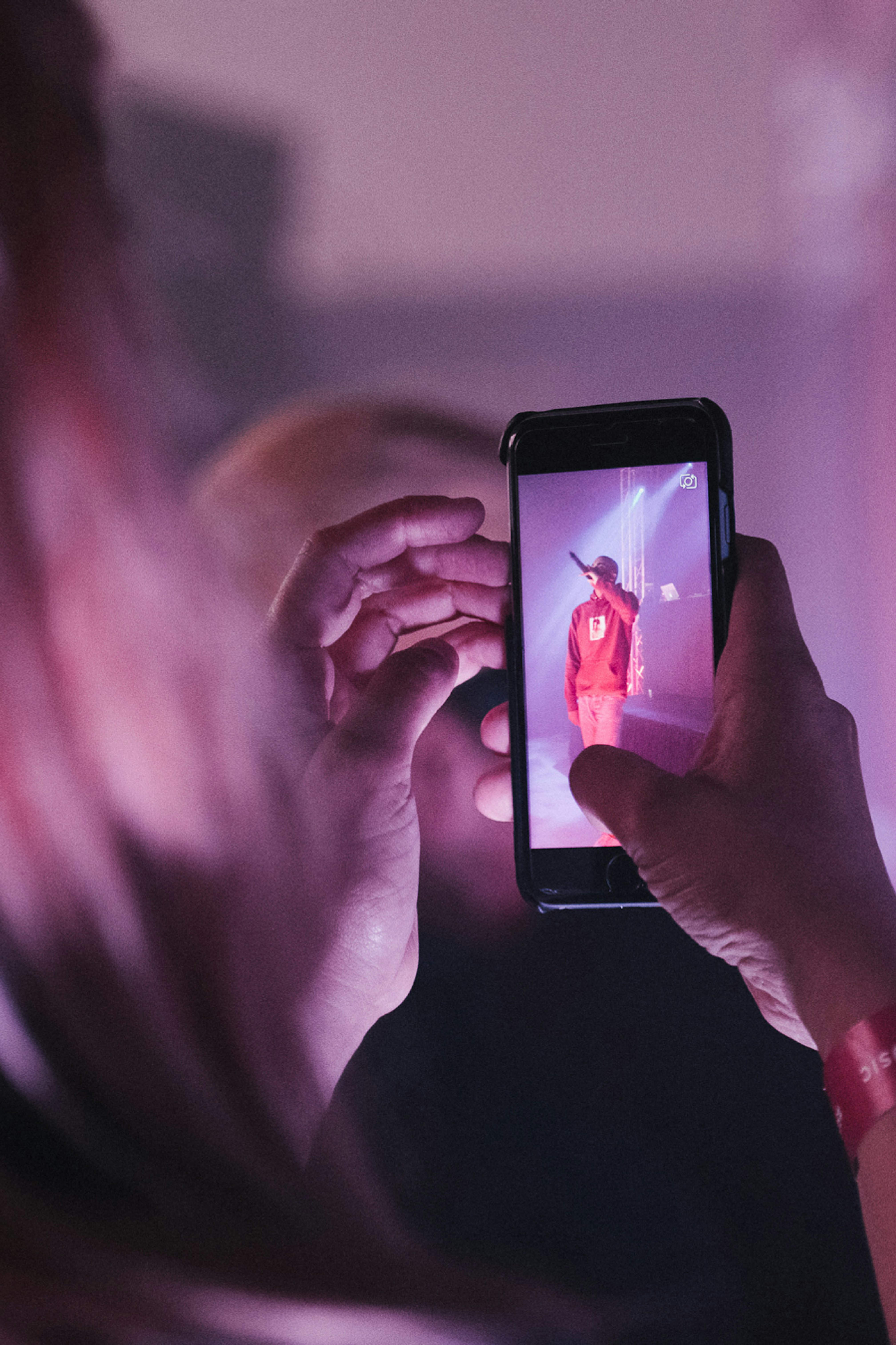 Image shows two hands holding a cell phone that is recording Vince Staples perform on stage with lights shining on him.