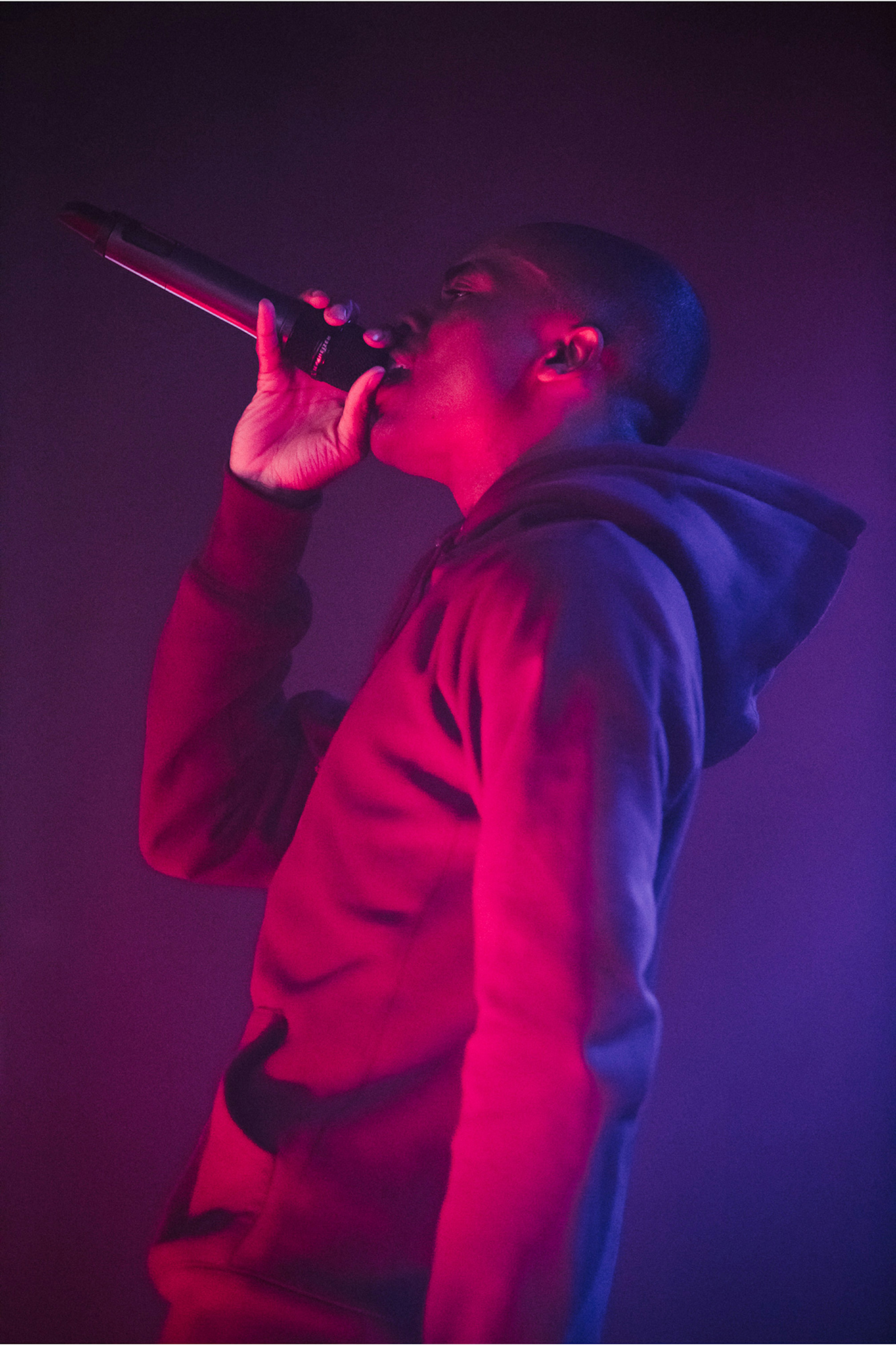 Image shows Vince Staples performing, holding a microphone to his face. He is wearing a hoodie, and there is a pink light shining on him. 