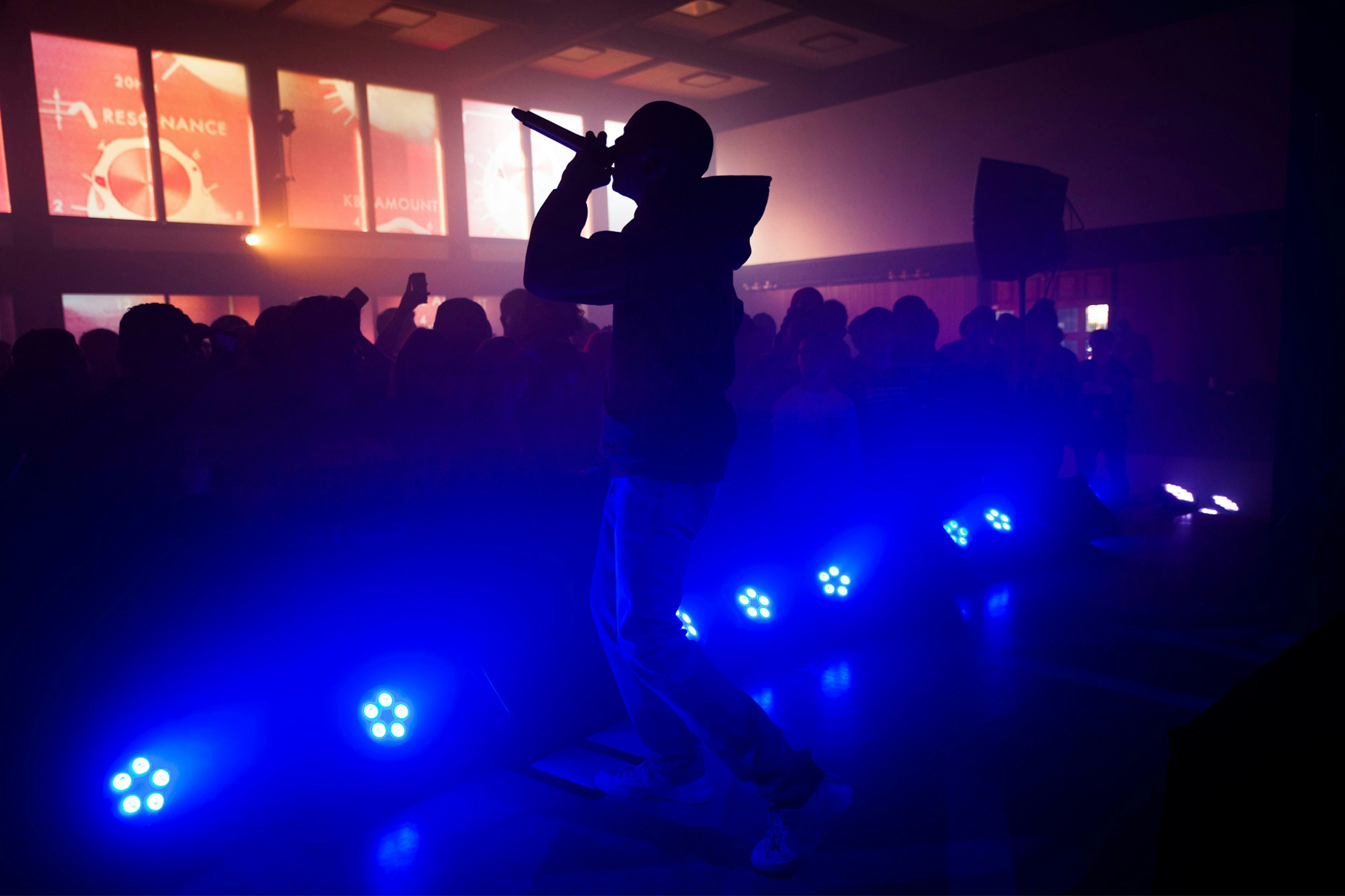 Image shows Vince Staples performing. He is silhouetted in front of a crowd and a row of blue lights shine on him. 